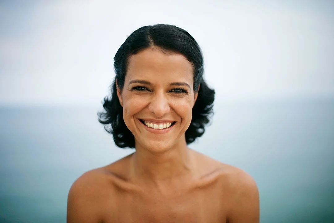 A white Brazilian woman and dark wavy hair smiling outdoors against a blurred background of water and clear sky at Ipanema beach in Rio de Janeiro, Brazil.