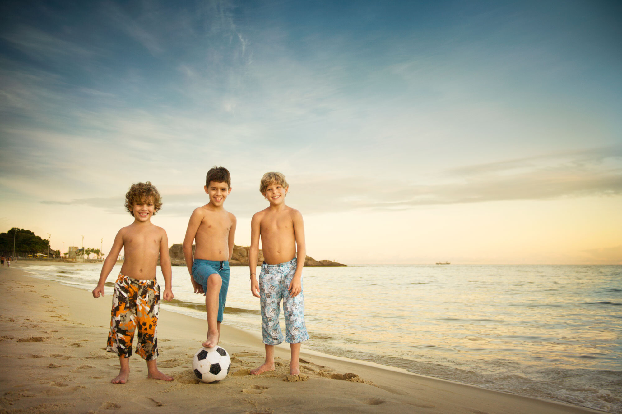 Three young Brazilian boys smiling on at Ipanema beach, with two of them standing and one balancing on a soccer ball, during sunset in Rio de Janeiro, Brazil.