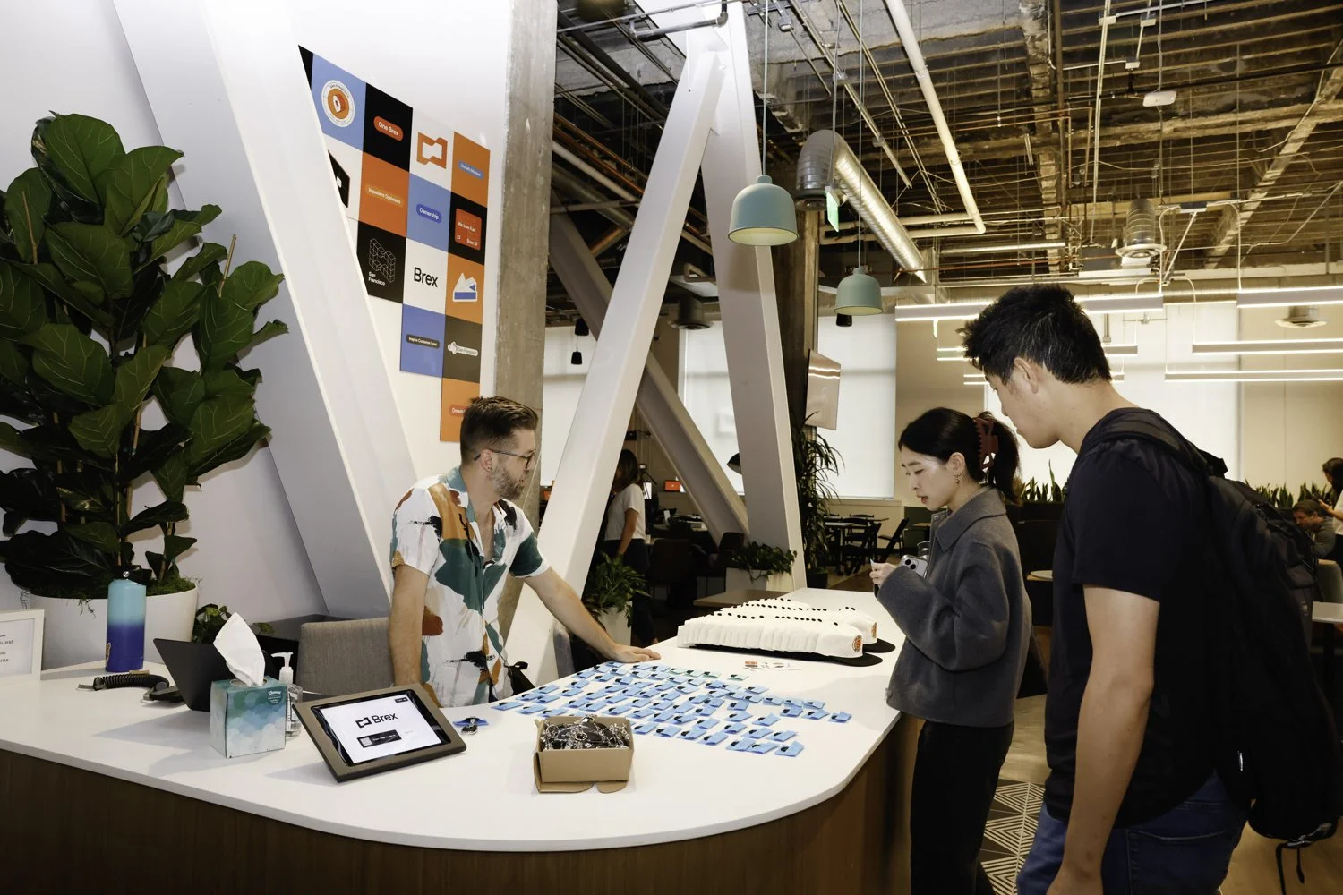A reception area where a woman is checking in at a modern office or coworking space. Two men are approaching the desk. The desk has small blue and white name tags or badges. Behind the desk, there's a man arranging the badges. The background features