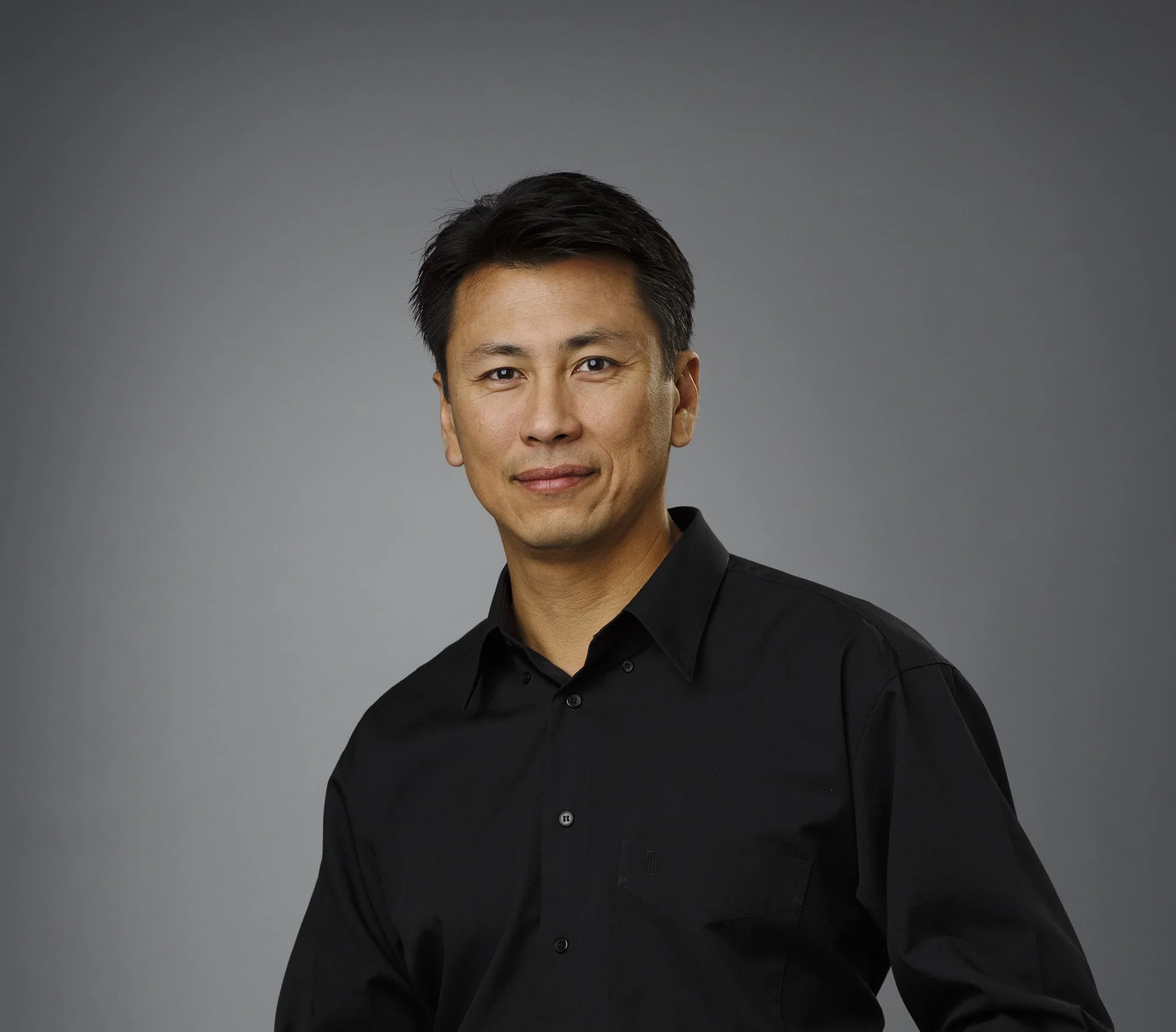 Portrait of an Asian man wearing a black button-up shirt, smiling slightly, against a plain gray background in San Francisco, CA.