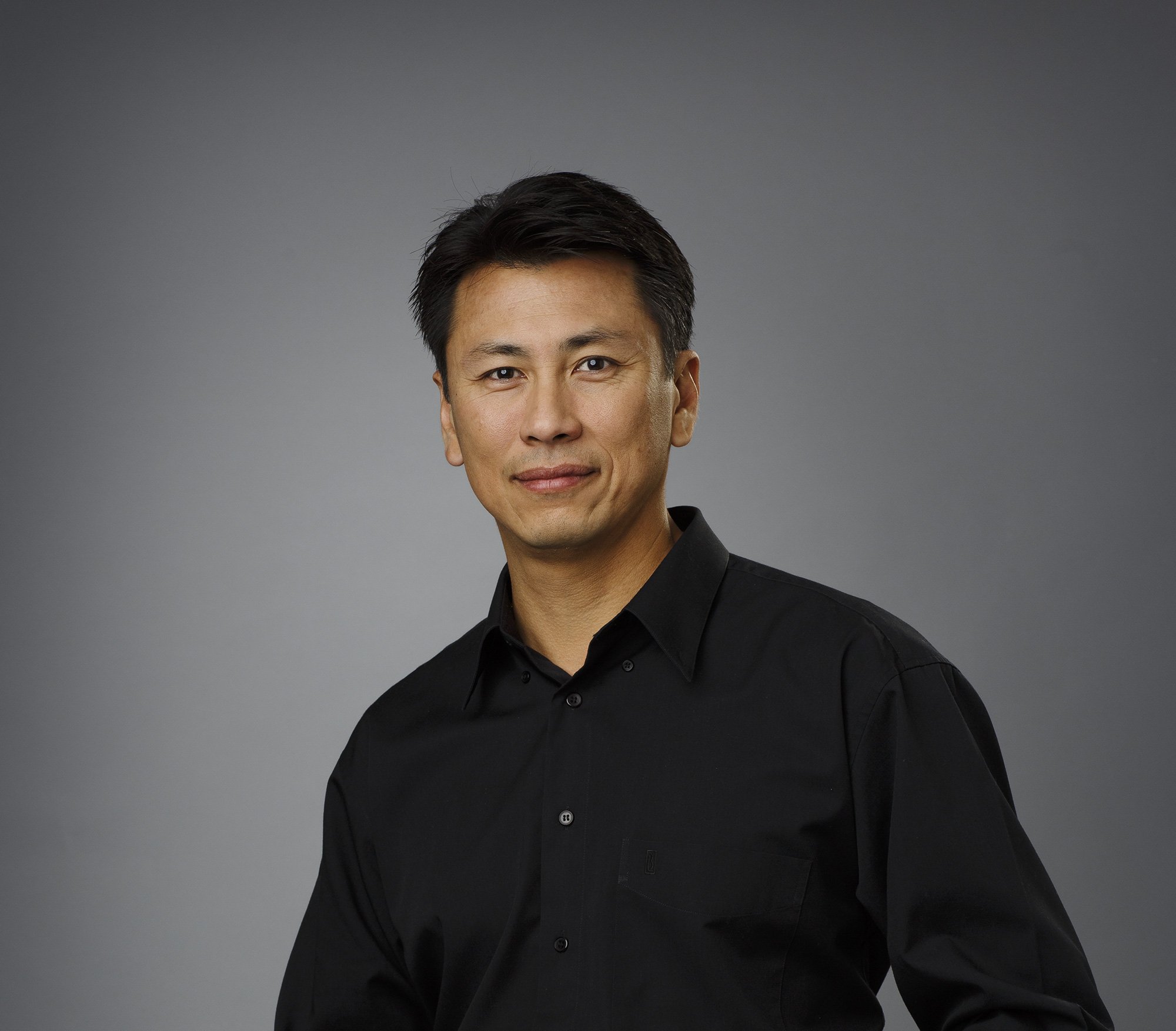 Portrait of an Asian man wearing a black button-up shirt, smiling slightly, against a plain gray background in San Francisco, CA.