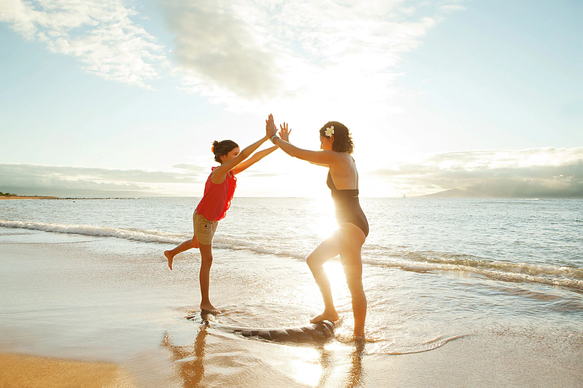 A Latina mother and a daughter are playing on the beach at sunset with backlit light, with the woman standing on a surfboard and both giving high fives in Maui, Hawai'i.