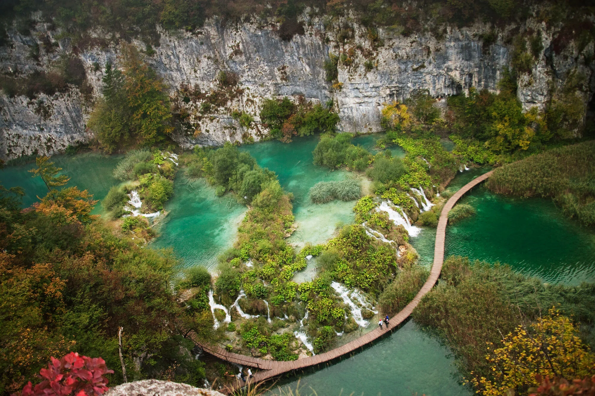Aerial view of a lush green landscape with waterfalls, turquoise lakes, and a winding wooden path in Croatia.
