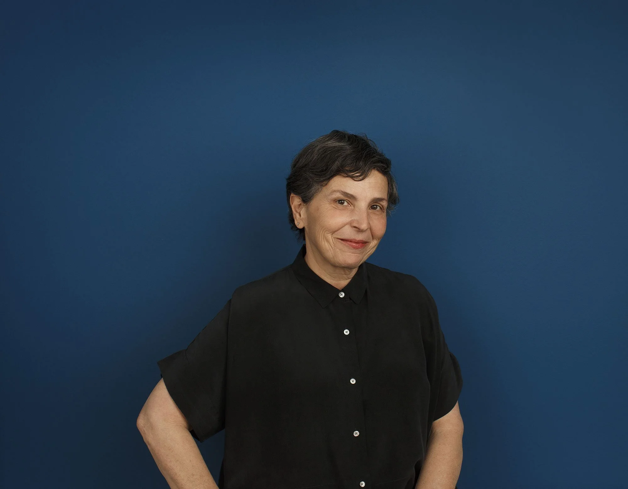 A Brazilian woman with short gray hair wearing a black button-up shirt, standing against a solid blue background, smiling softly with her left hand on her hip at an architectural office in San Francisco, CA.