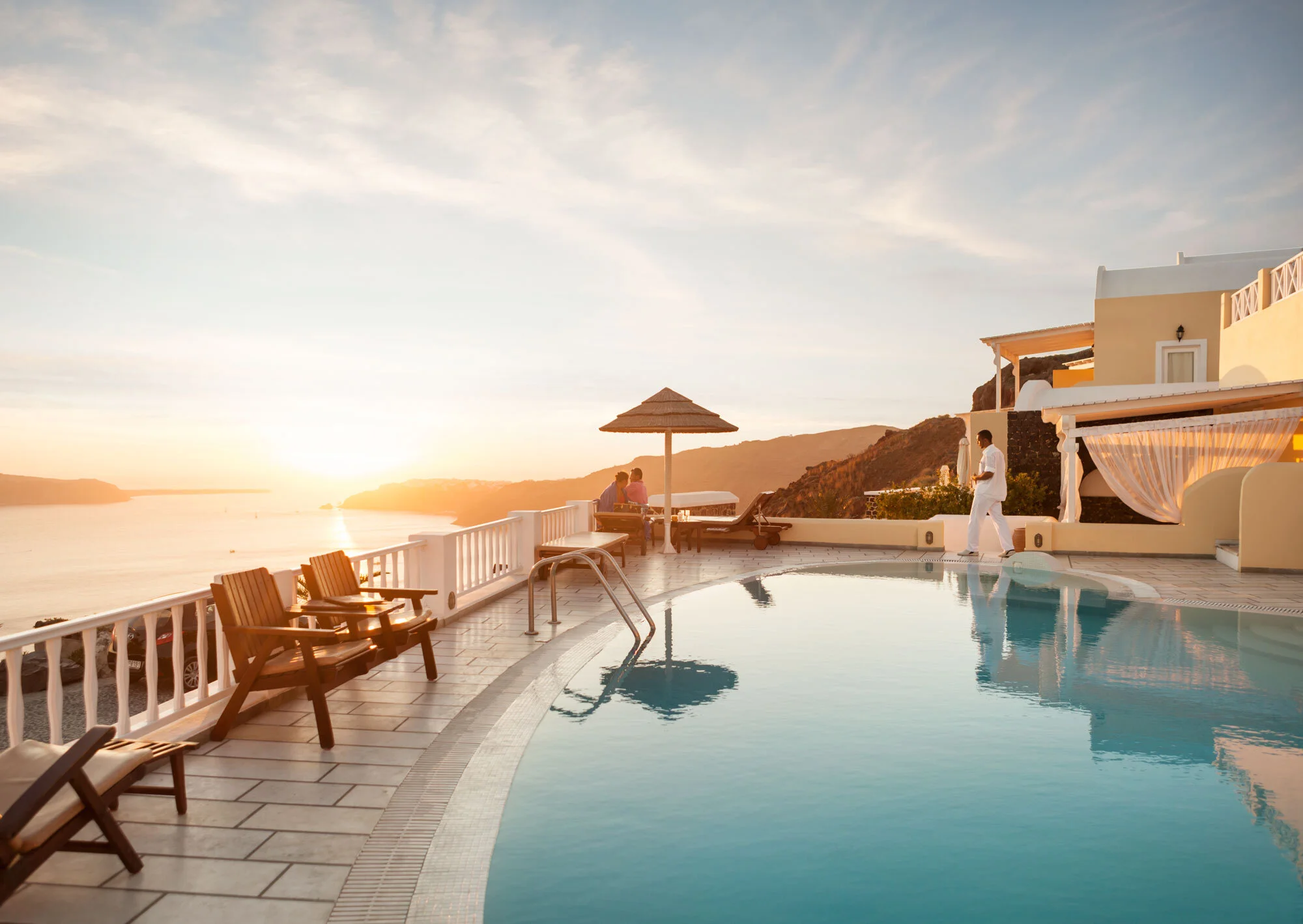 A luxurious infinity pool overlooking the ocean at sunset, with lounge chairs, a couple sitting under a straw umbrella, and a person in white attire walking nearby, in a coastal resort setting in Santorini, Greece.