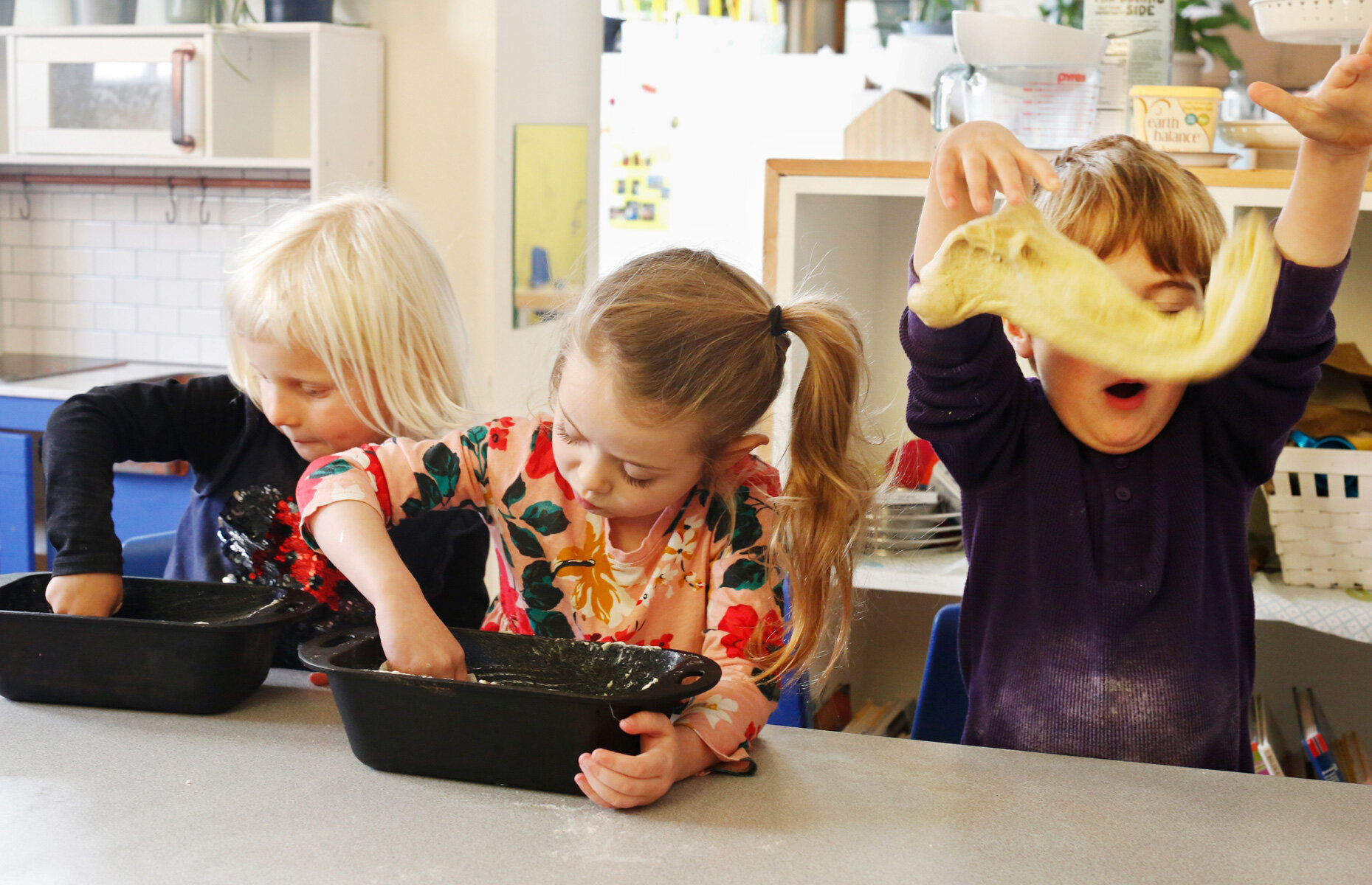 Three children with blonde and red hair engaged in a cooking activity at a table. One boy is holding and stretching dough above his head, with dough covering his face. Two girls are buttering baking pans at the The Scandinavian School in San Francisc
