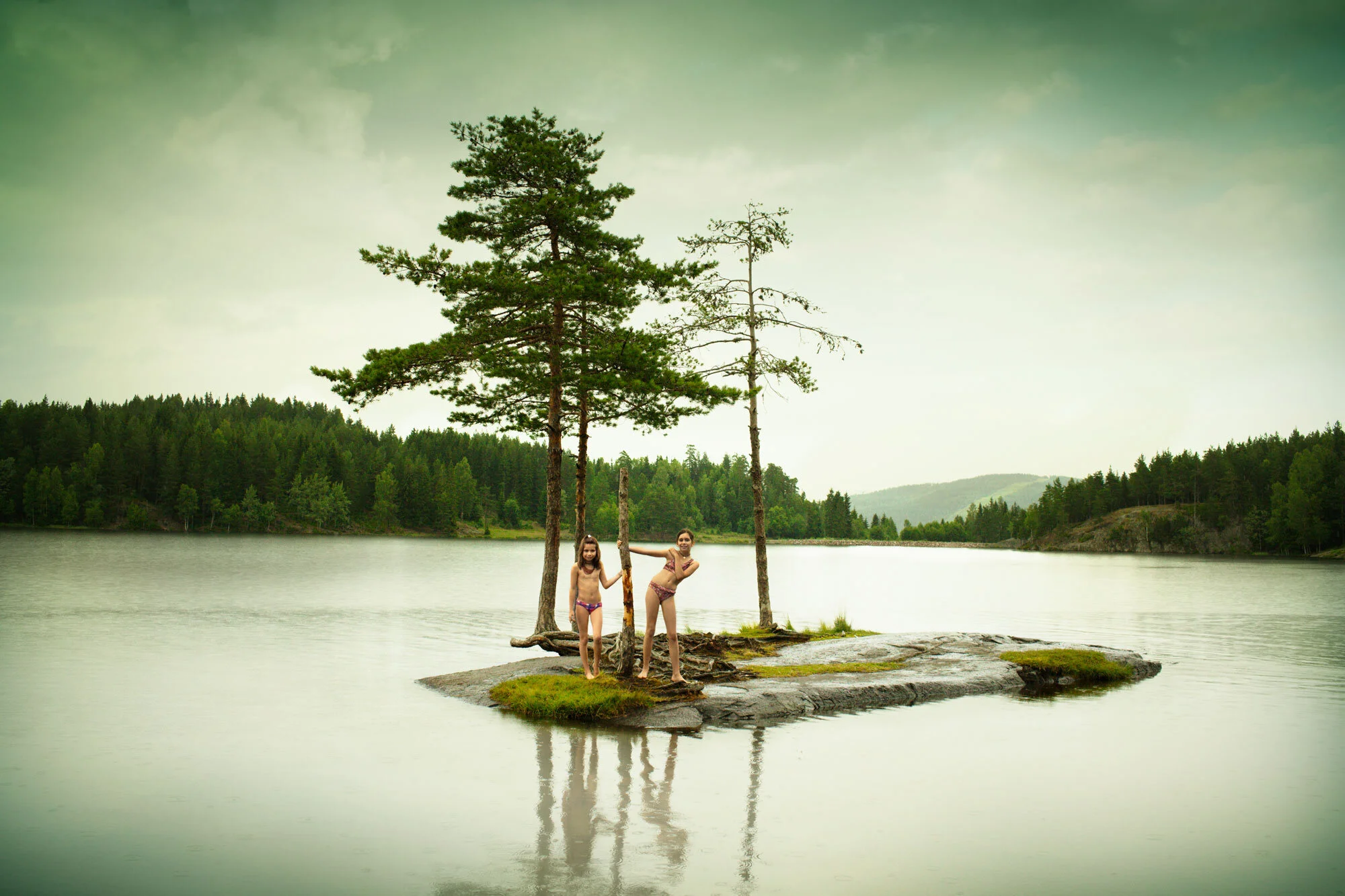 Two Norwegian children in swimsuits standing on a small island with trees, surrounded by a lake and a forested landscape in the background in Oslo, Norway.