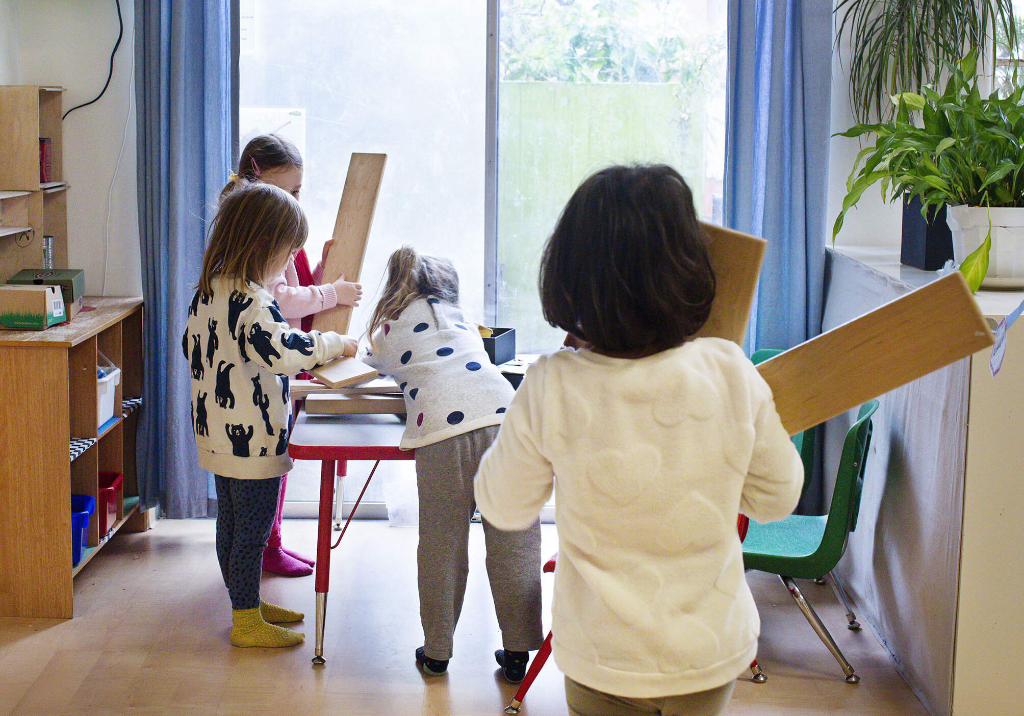 Four children in a classroom, each holding wooden boards, engaged in an activity near a large window with blue curtains and a view of greenery outside. A plant is on a windowsill at the The Scandinavian School in San Francisco, CA.