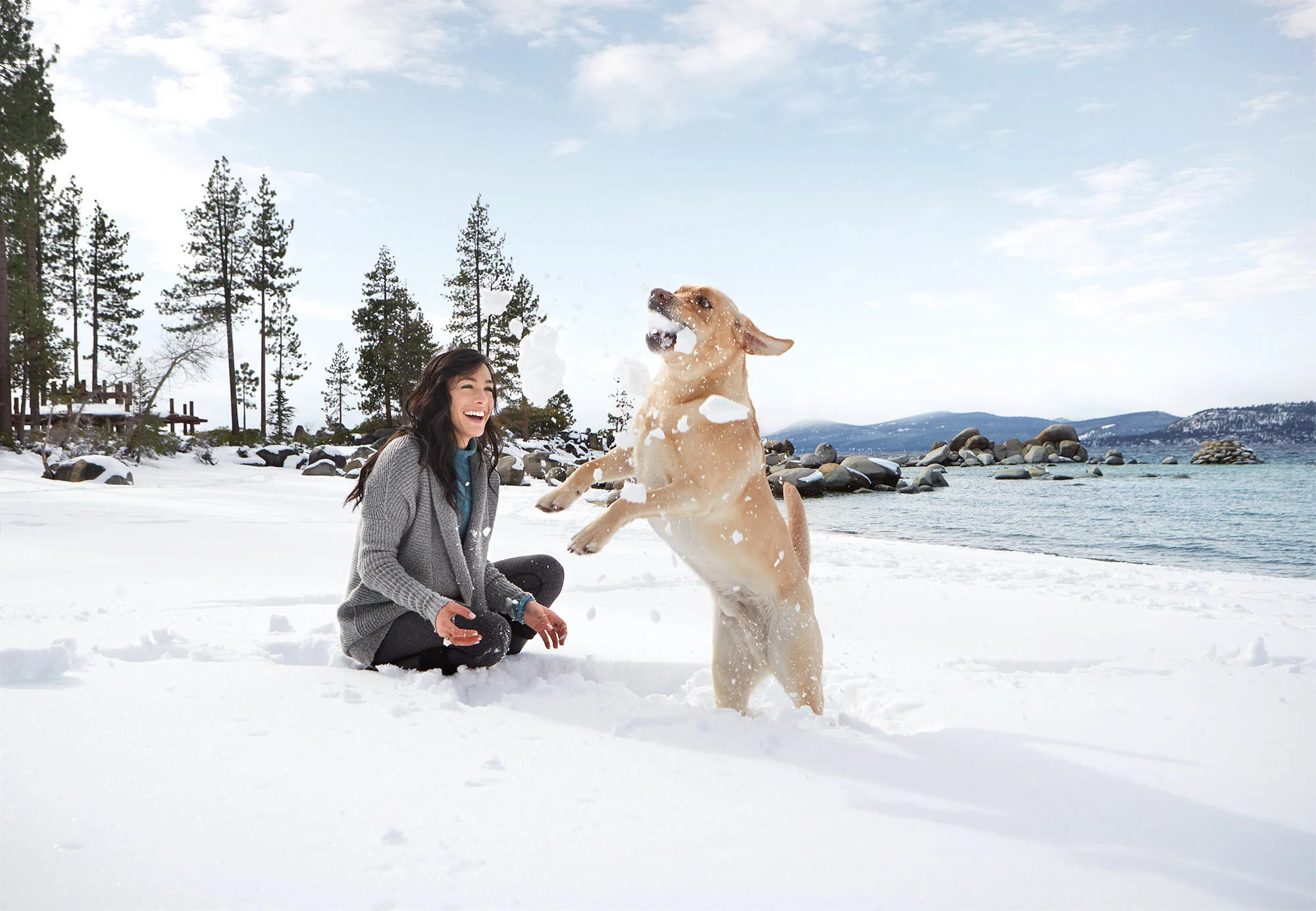A woman with long black hair and a dog playing in the snow by a lake, with trees and mountains in the background against a blue sky with clouds at Sand Harbor, Lake Tahoe, CA.