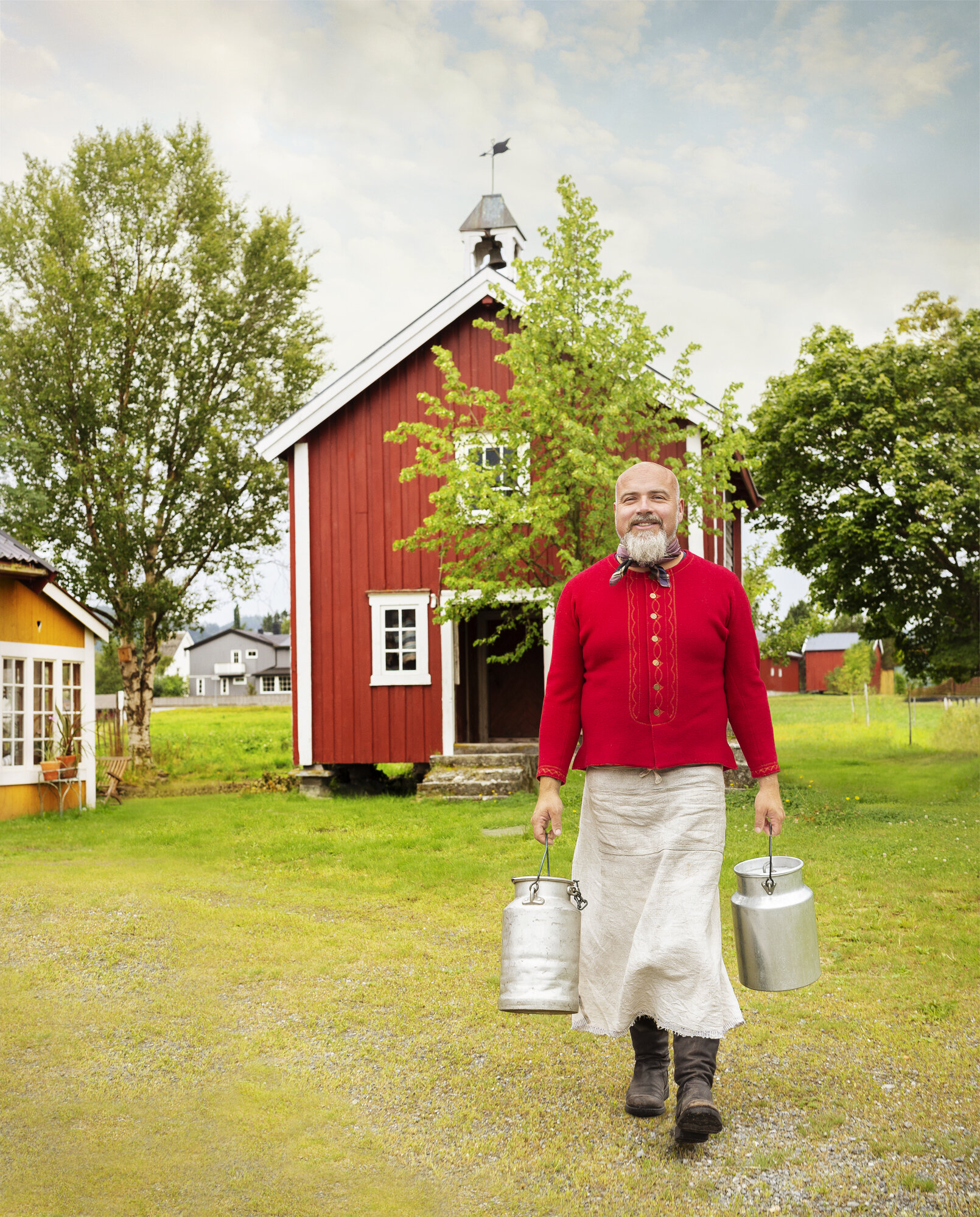 A white Norwegian farmer with a bald head, beard wearing a red sweater, apron, and brown boots walking on grass carrying two milk cans in his hands, in front of a red barn and green trees in Norway.