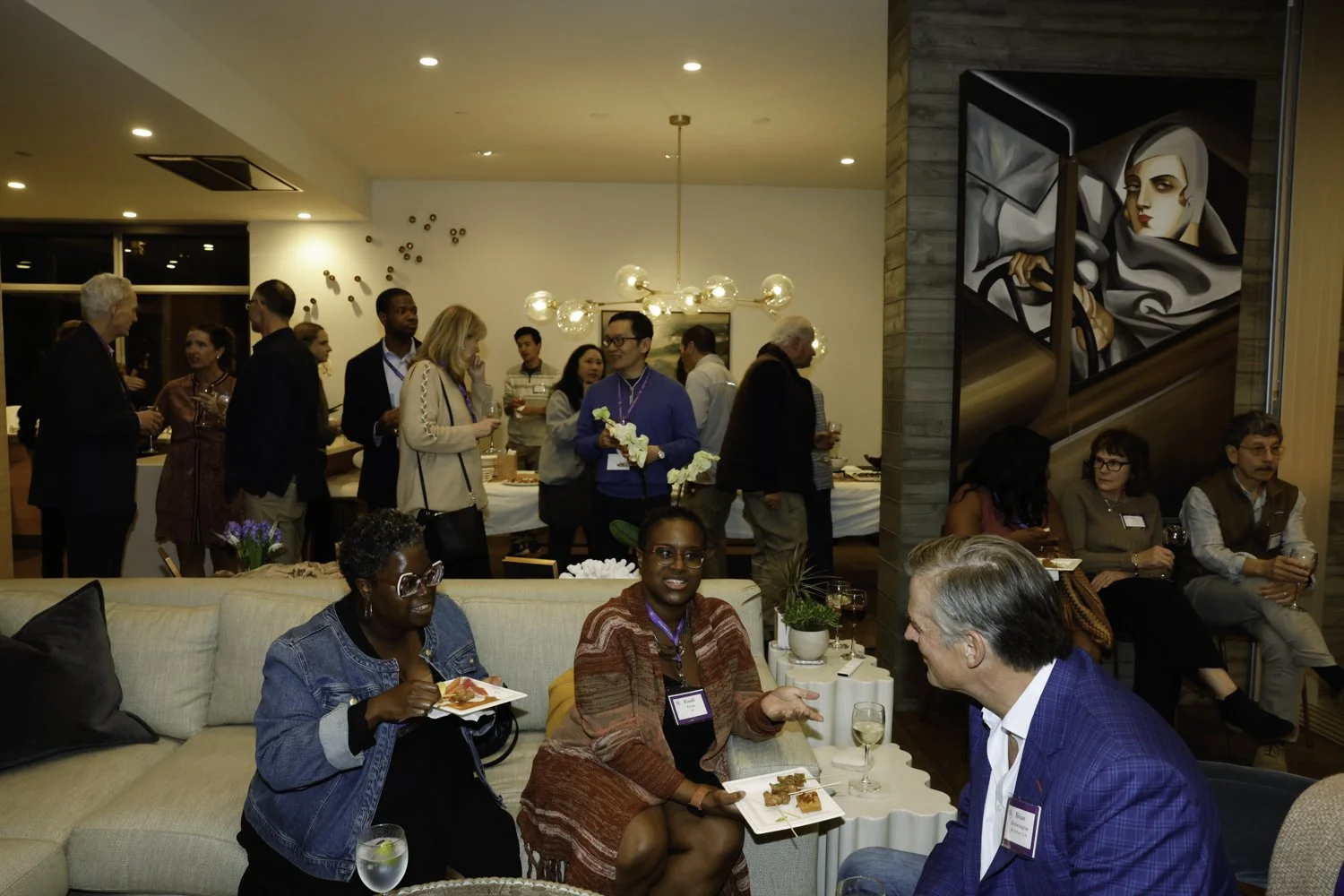 A group of diverse people attending a social networking event in a warmly lit room. Some are standing and talking in the background, while others are seated on a beige couch and chairs, enjoying snacks and drinks. There is modern artwork on the walls