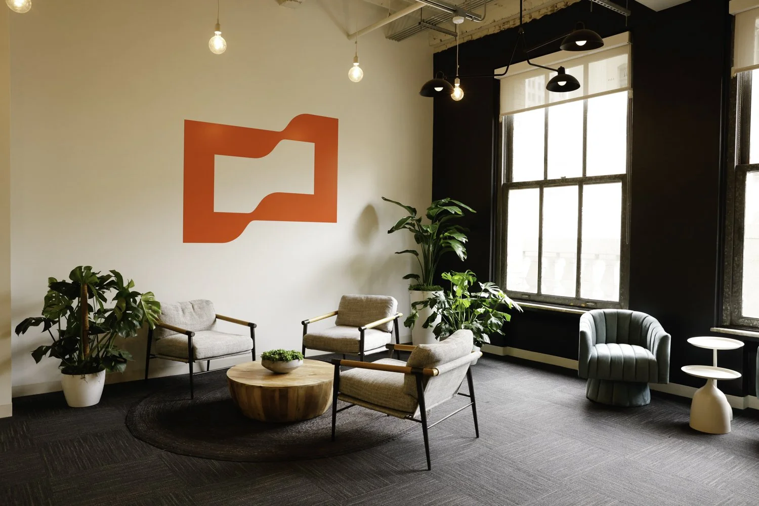 A modern office waiting area with three light-colored armchairs around a round wooden coffee table, several potted plants, and a black accent wall with a red geometric artwork. Large windows provide natural light, and black pendant lamps hang from th