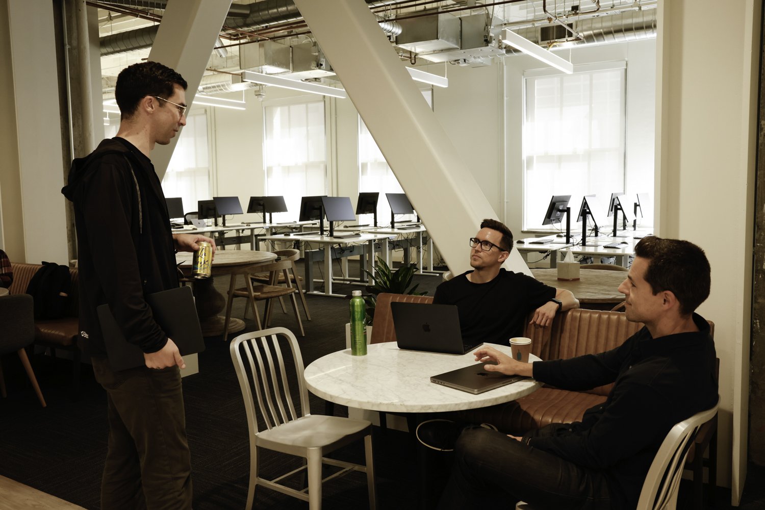 Three men having a conversation in an office lounge area with computers in the background. Two men are seated at a round table with laptops, and one man is standing holding a drink at Brex Headquarter in San Francisco, CA.