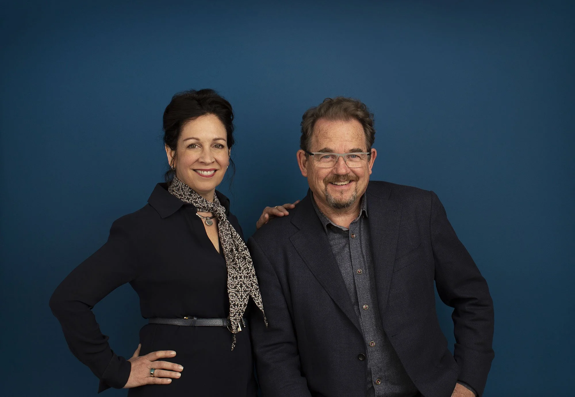 A white couple smiling together against a blue background. The woman has dark hair styled up, wearing a black blazer and a patterned scar. The man has glasses and a beard, wearing a dark blazer over a dark shirt in San Francisco, CA.