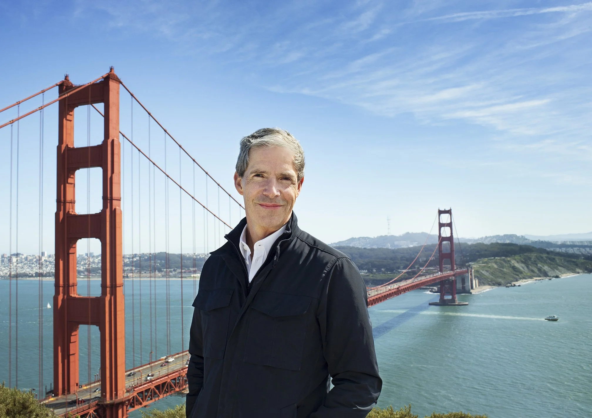 Greg Flynn with the Golden Gate Bridge in the background on a sunny afternoon in San Francisco, CA. Greg is wearing a black jacket and a white shirt. The water is green-blue behind him and the sky is also blue with wispy clouds.