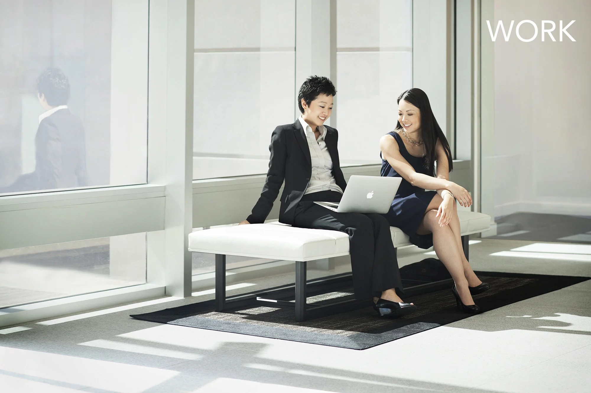 Two women sitting on a bench in an office lobby, smiling and looking at an Apple laptop. One woman is wearing a black suit, the other a navy dress. The background shows large glass windows and a wall with the word 'WORK' written on it.