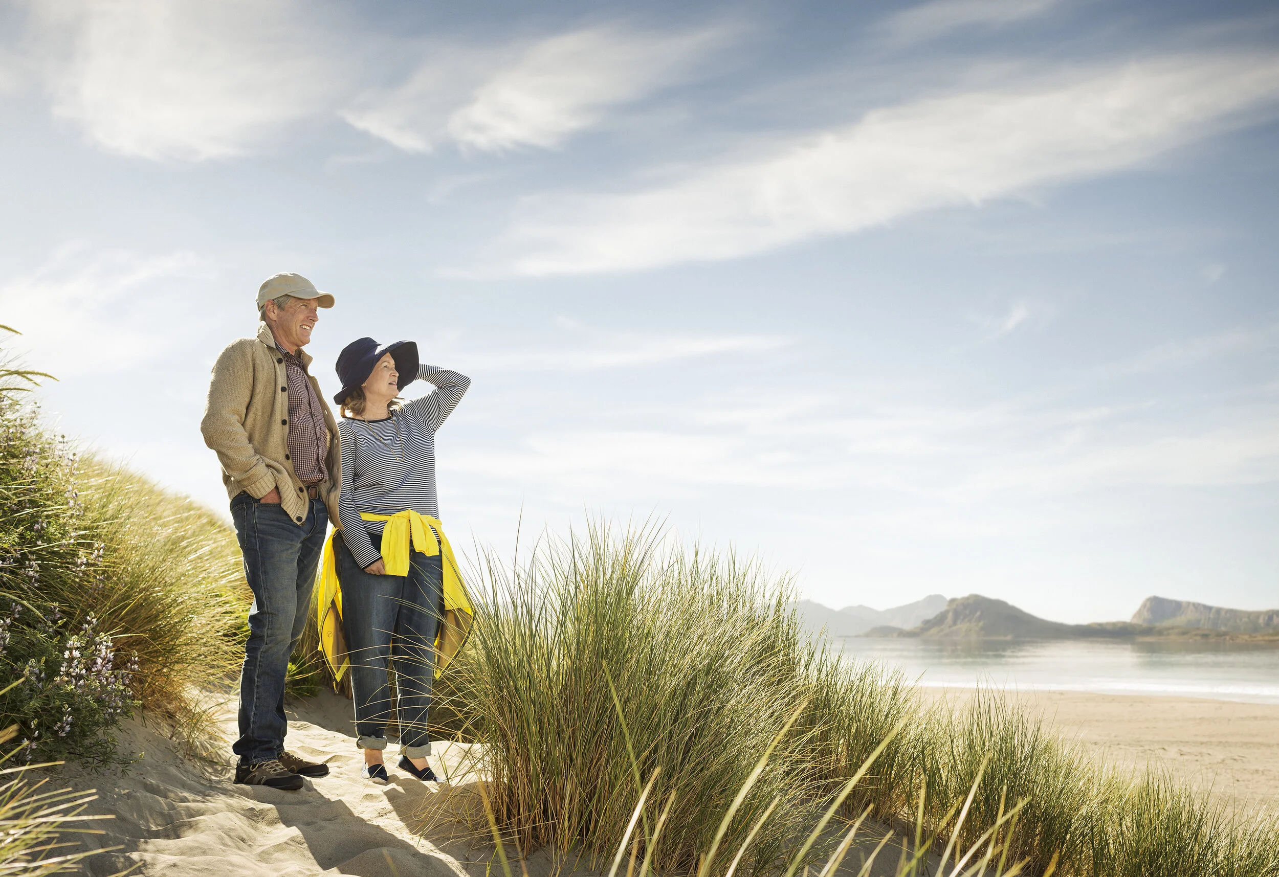 A white middle aged couple, man and woman stand on a sandy beach with tall grasses, looking out across the ocean and distant mountains on a clear day for Genentech Pharma, Wellness at Limantour beach, CA.