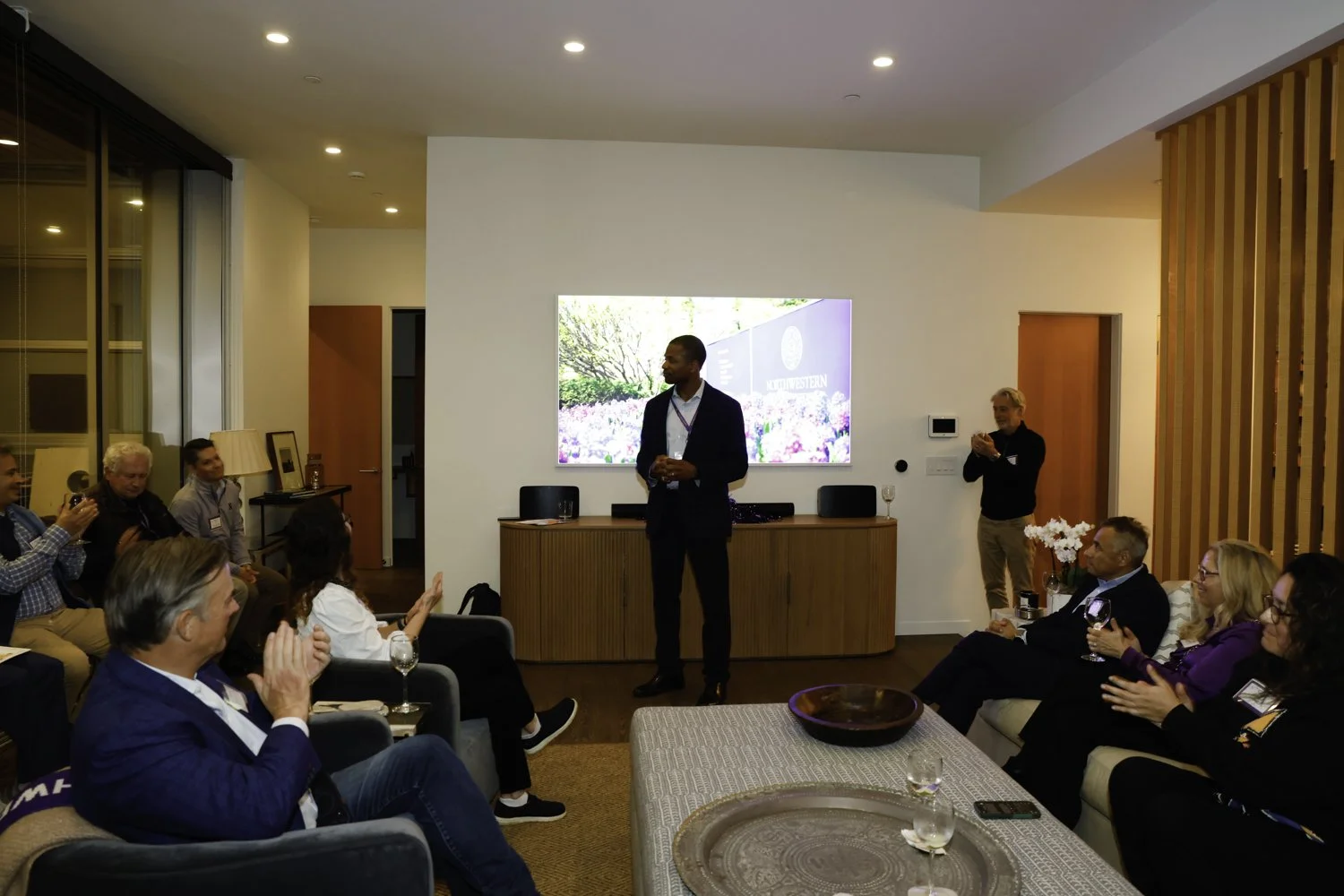 A man in a suit standing in front of an audience in a modern living room, giving a presentation or speech. The audience is seated on couches and chairs, applauding and listening attentively. There is a large screen on the wall behind the speaker disp