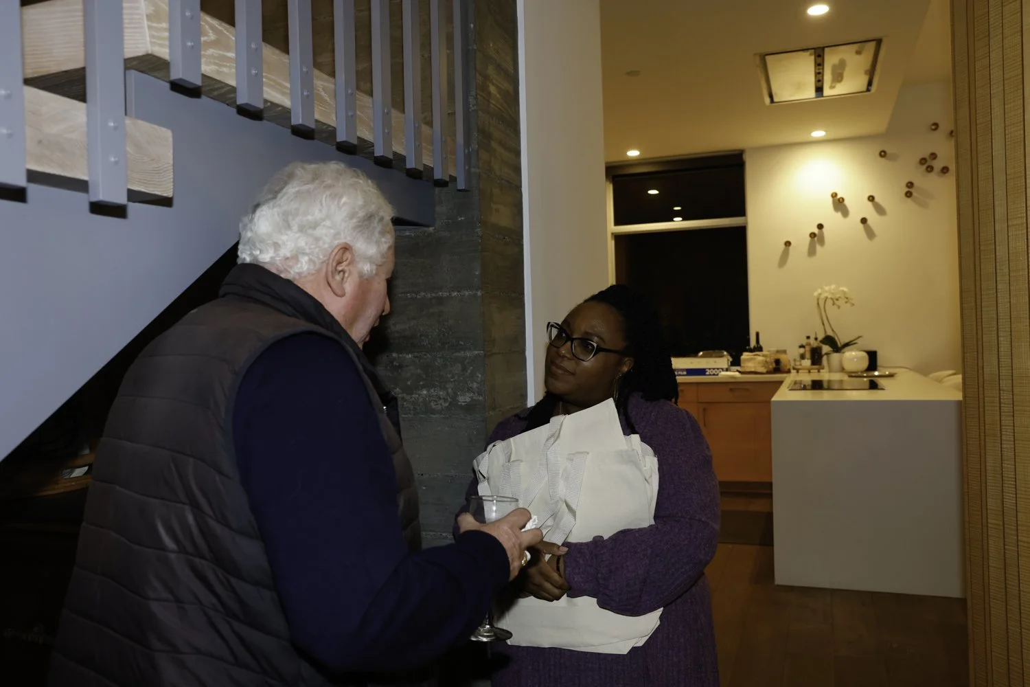Two people talking at a gathering in a home. An older man with white hair holds a glass and is engaged in conversation with a woman wearing glasses and a purple sweater. The woman holds a white package or bag at Northwestern fundraiser in San Francis
