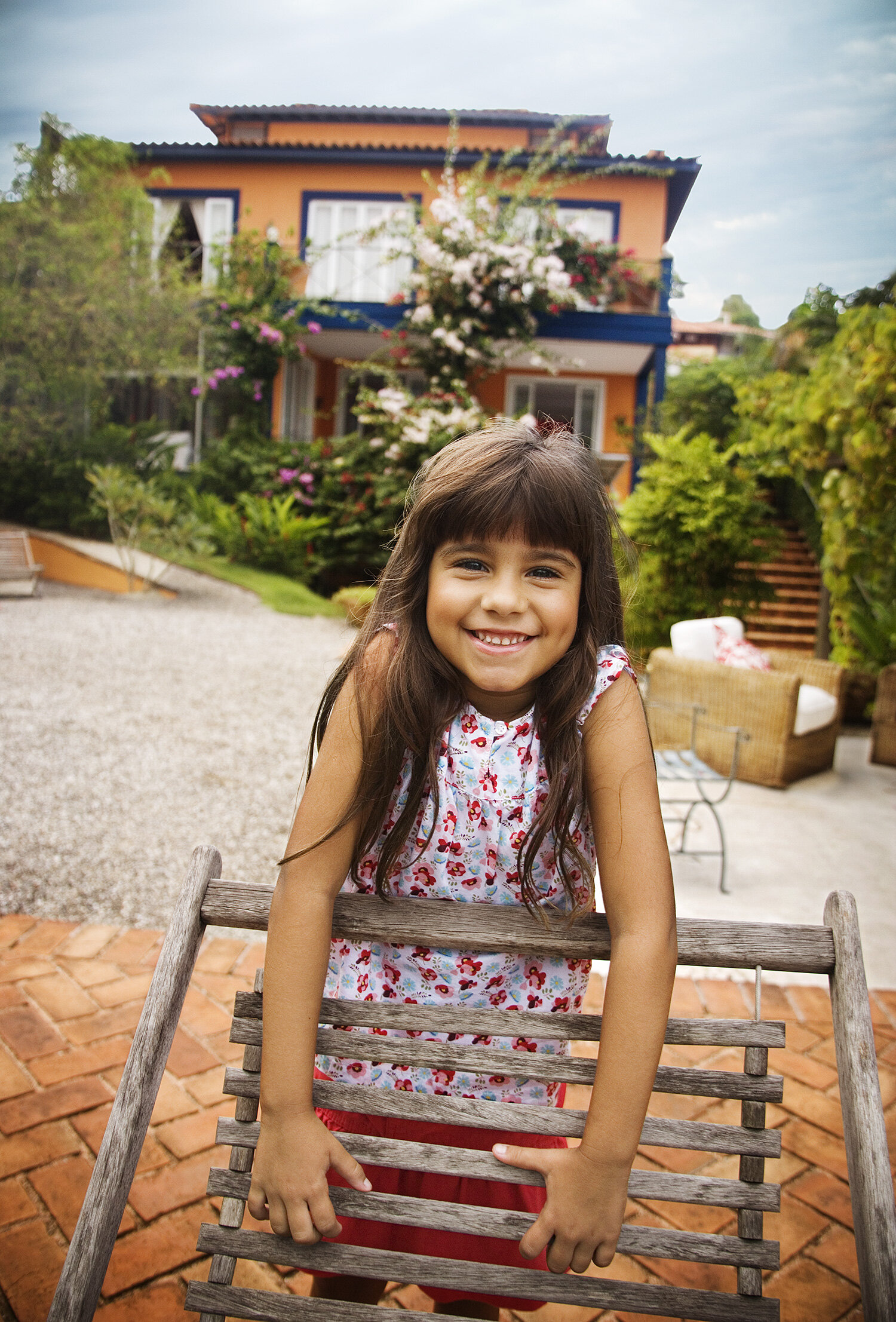 A happy young Brazilian girl with long brown hair, wearing a white dress with red and pink flowers, smiling and sitting behind a rustic wooden chair in a backyard setting with a house, trees, and cloudy sky in the background in Rio de Janeiro, Brazil