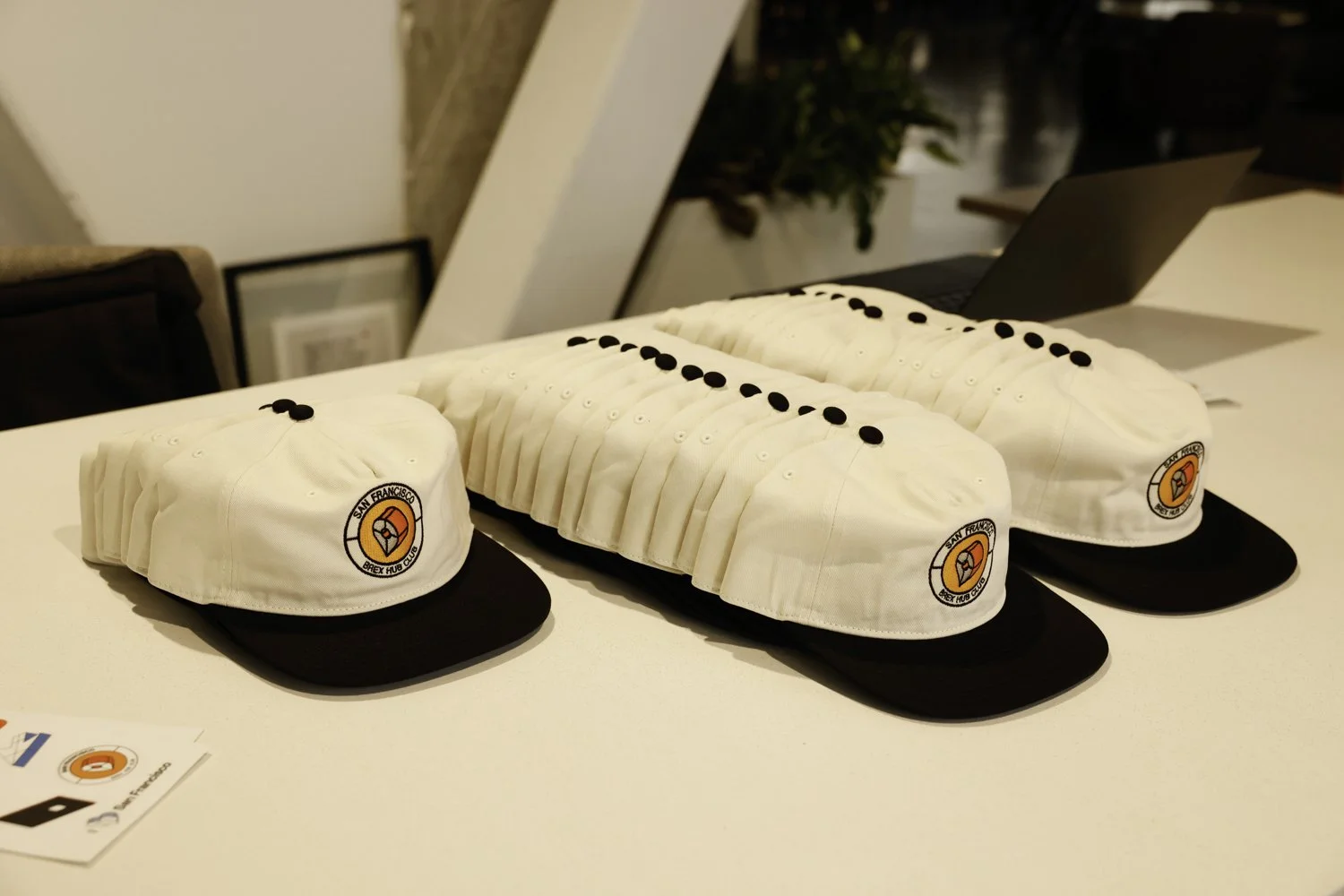 Four white baseball hats with black brims and the San Francisco Brex Hub Club logo placed on a table, with some wearing black buttons and others with black stickers on the hats Brex headquarter in San Francisco, CA.
