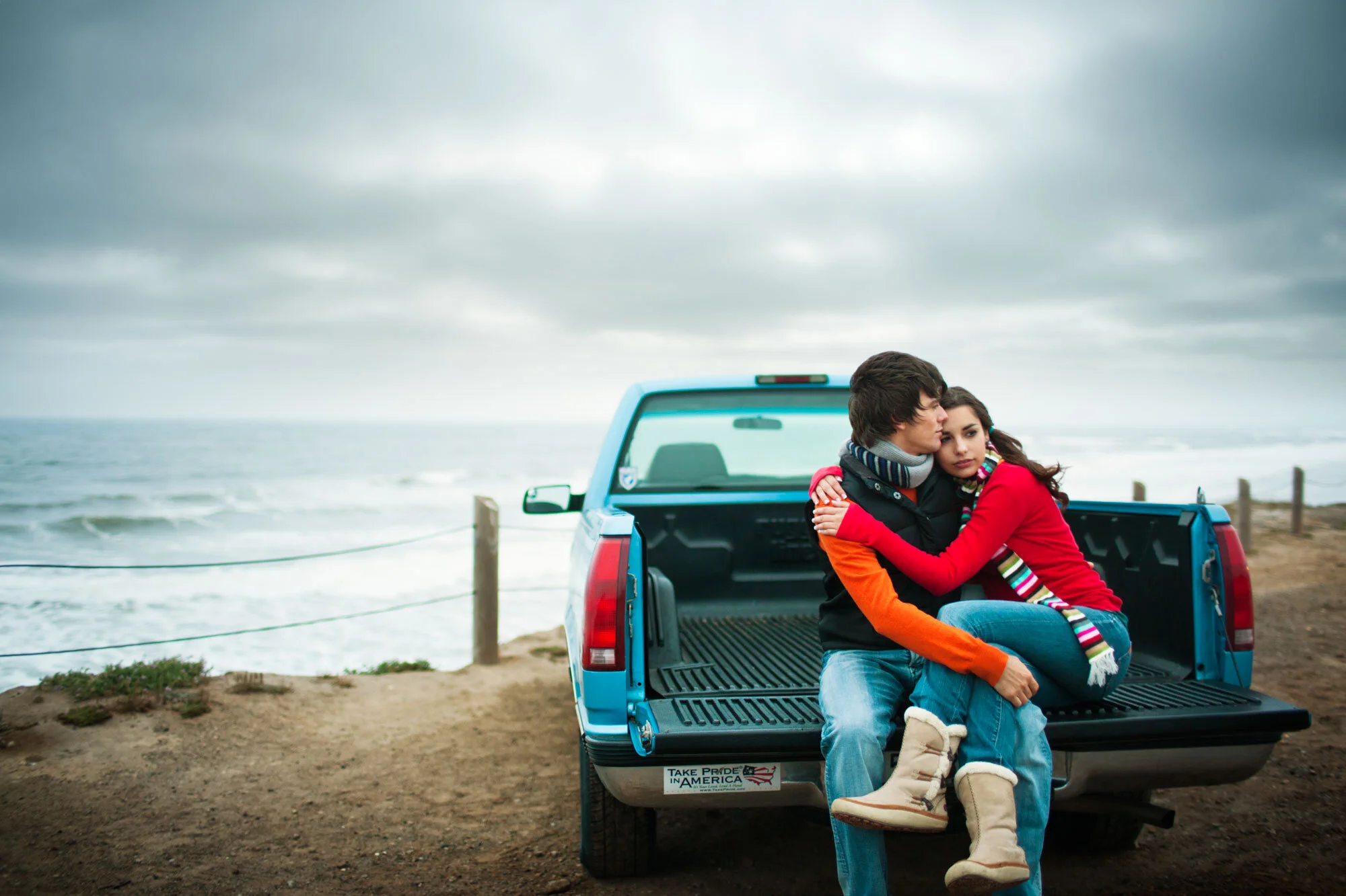Young Latino couple sitting on the back of a blue pickup truck parked near the ocean on a cloudy day, embracing each other with the ocean and cloudy sky in the background at Ocean Beach in San Francisco CA.