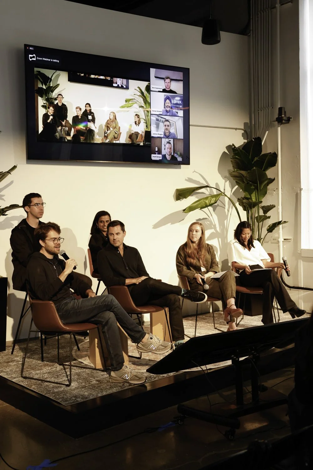 Panel discussion with five seated individuals, one holding a microphone, on a stage with large plants and a large screen displaying a Zoom call with multiple participants Brex headquarter in San Francisco, CA.