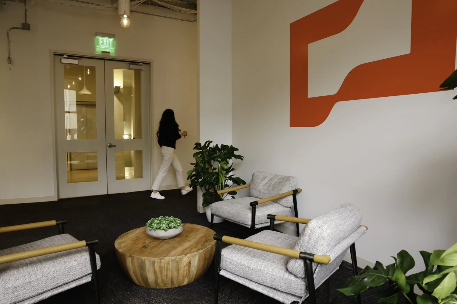 A woman walking towards a glass door in a modern, minimalistic lounge area with gray armchairs, a wooden coffee table with a green plant, and modern wall art. The space has natural light and indoor plants Brex headquarter in San Francisco, CA.