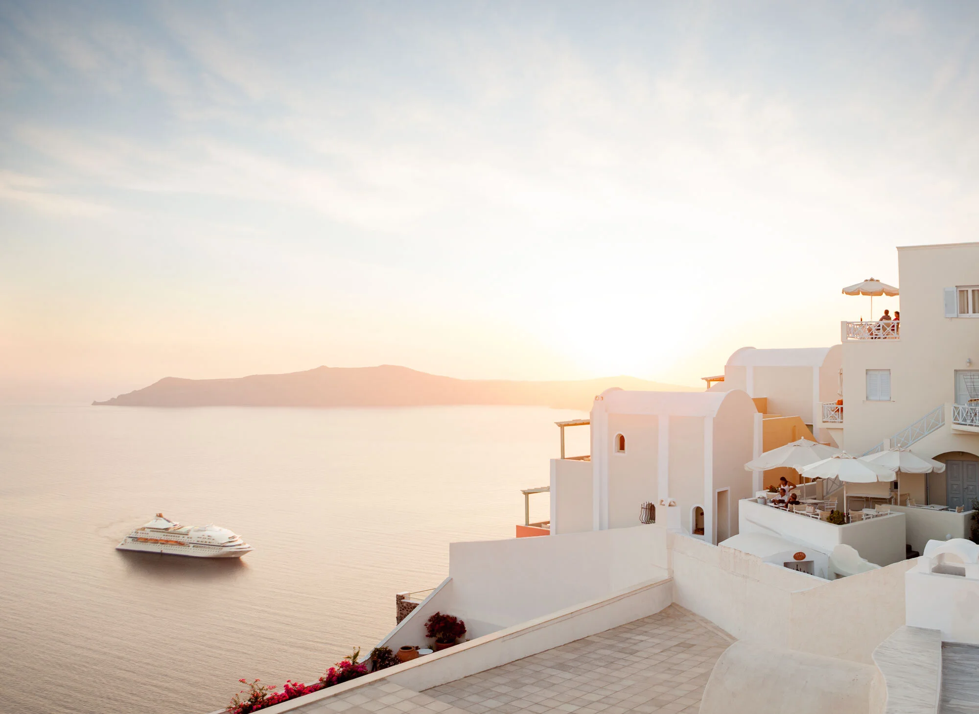 White Mediterranean-style buildings with balconies and outdoor seating overlooking the sea at sunset, with a large cruise ship passing by in the water.