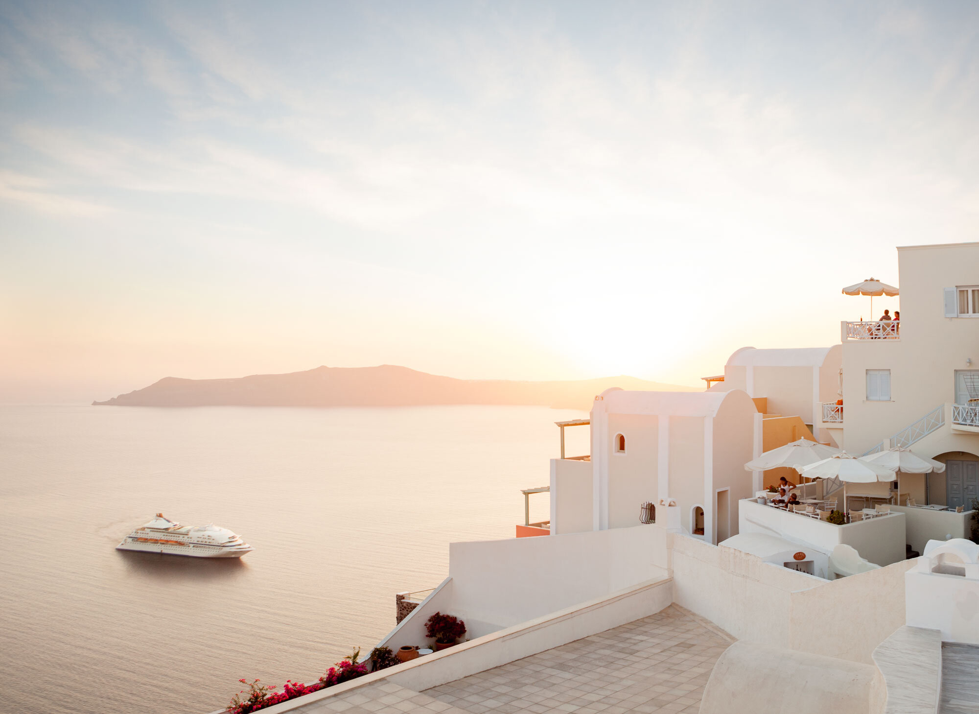White Mediterranean-style buildings with balconies and outdoor seating overlooking the sea at sunset, with a large cruise ship passing by in the water in Santorini, Greece.