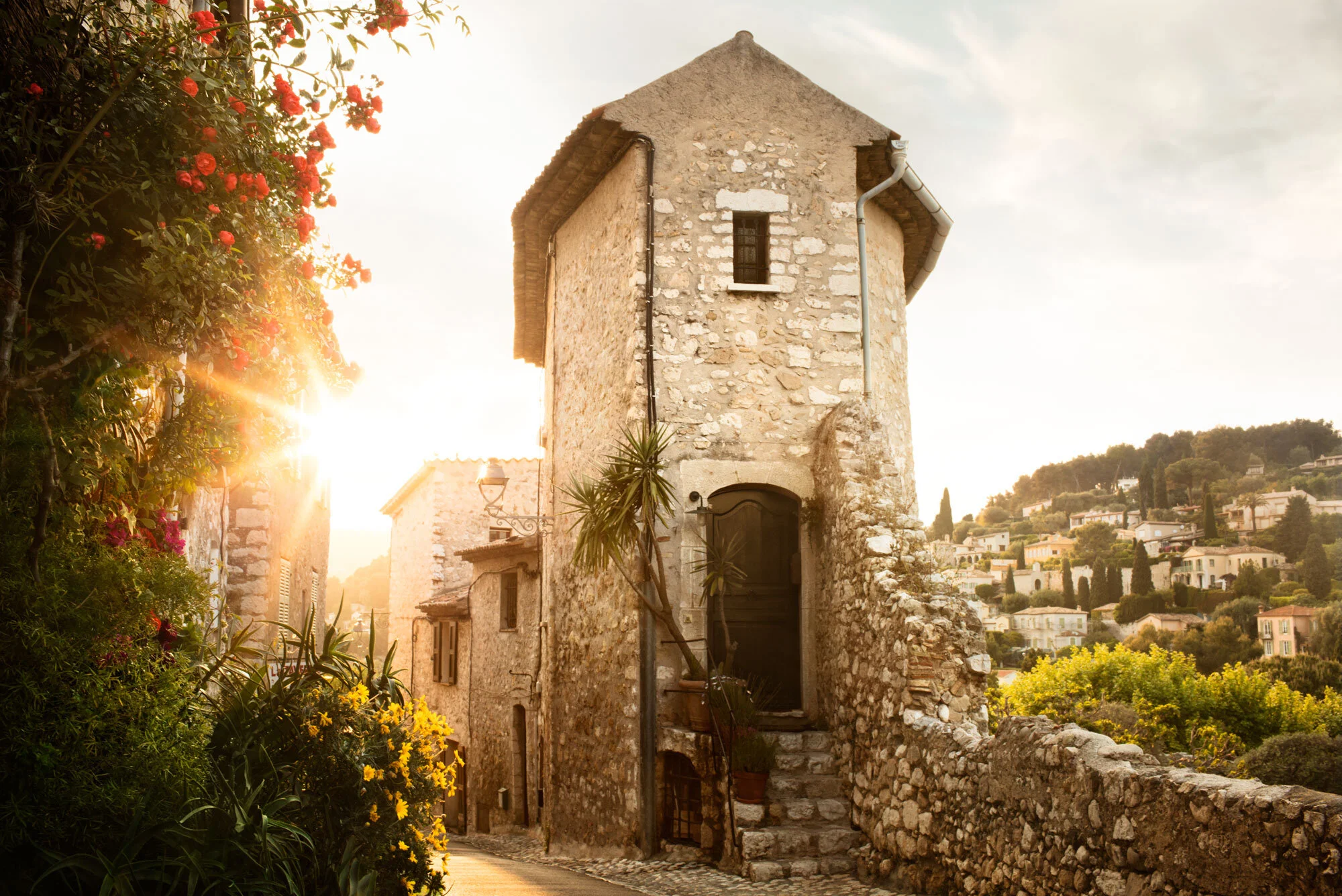 A rustic stone house with a stepped staircase in a hillside village at sunset in Saint-Paul de Vence, France, surrounded by vibrant flowering plants and overlooking a landscape of houses and trees.