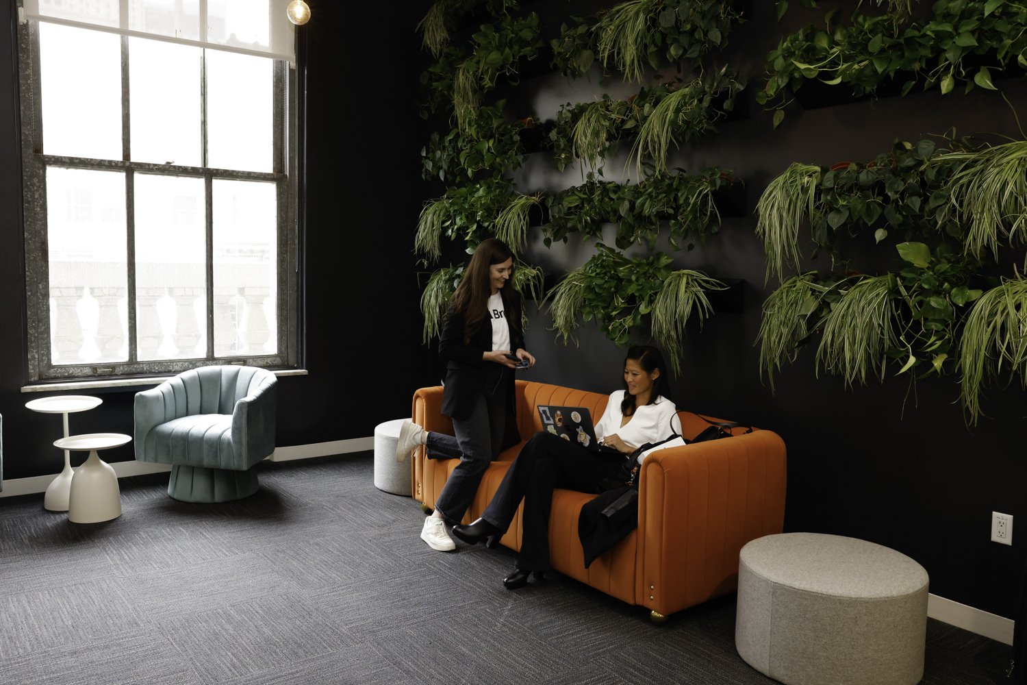 Two women in a modern lounge area with a large window, green wall of plants, orange sofa, mint green chair, and white tables, engaging in conversation Brex headquarter in San Francisco, CA.