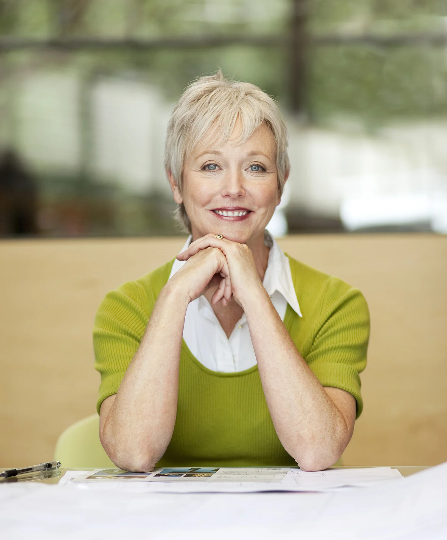 A smiling white middle-aged woman with short white hair, wearing a white blouse and bright green sweater, sitting at a table with papers, in an architectural office in San Francisco, CA.