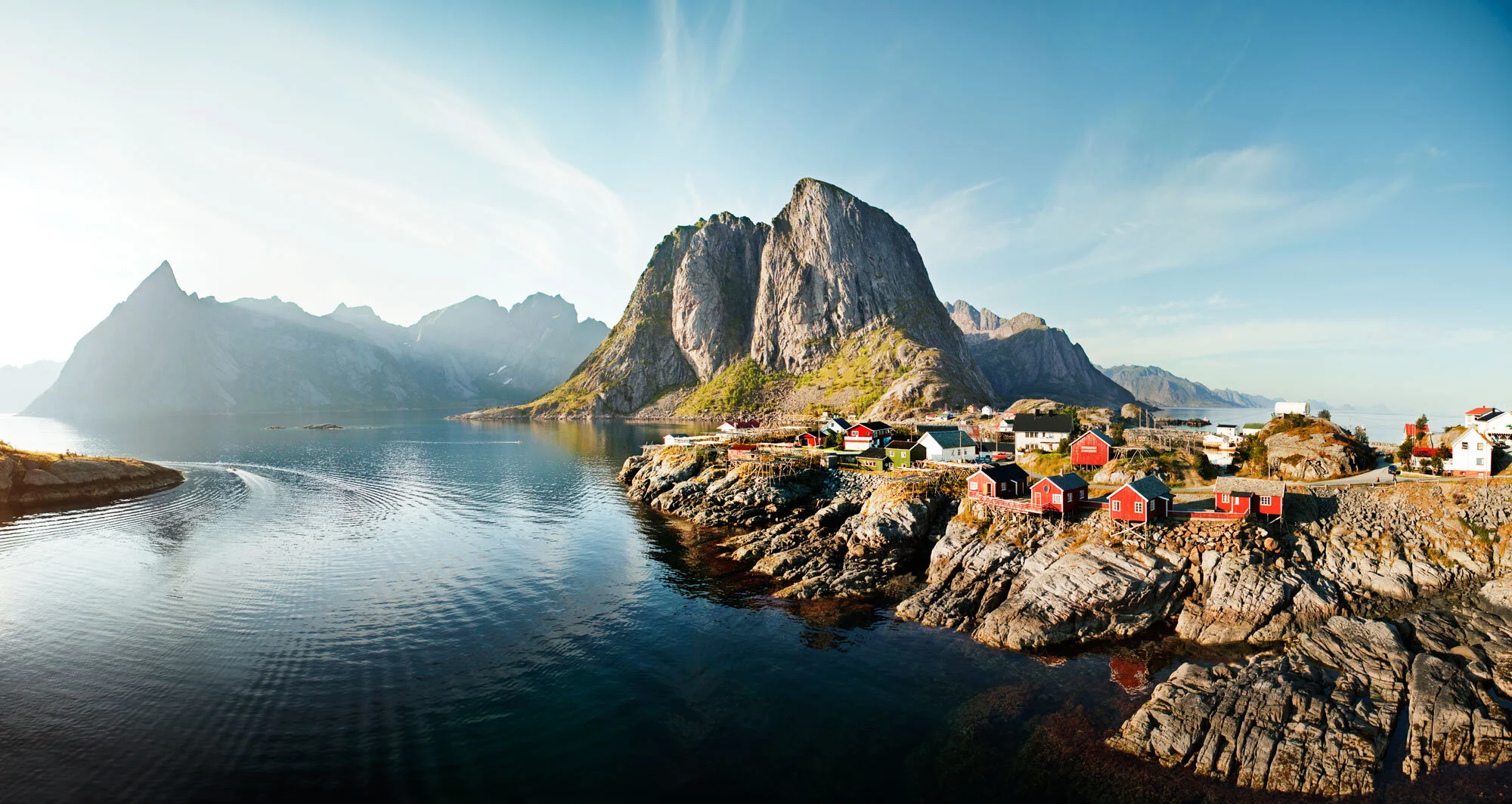 A scenic coastal village with colorful fishermen's cabins on rocky cliffs, a calm water body, and tall mountains in the background under a blue sky in Lofoten, Norway.