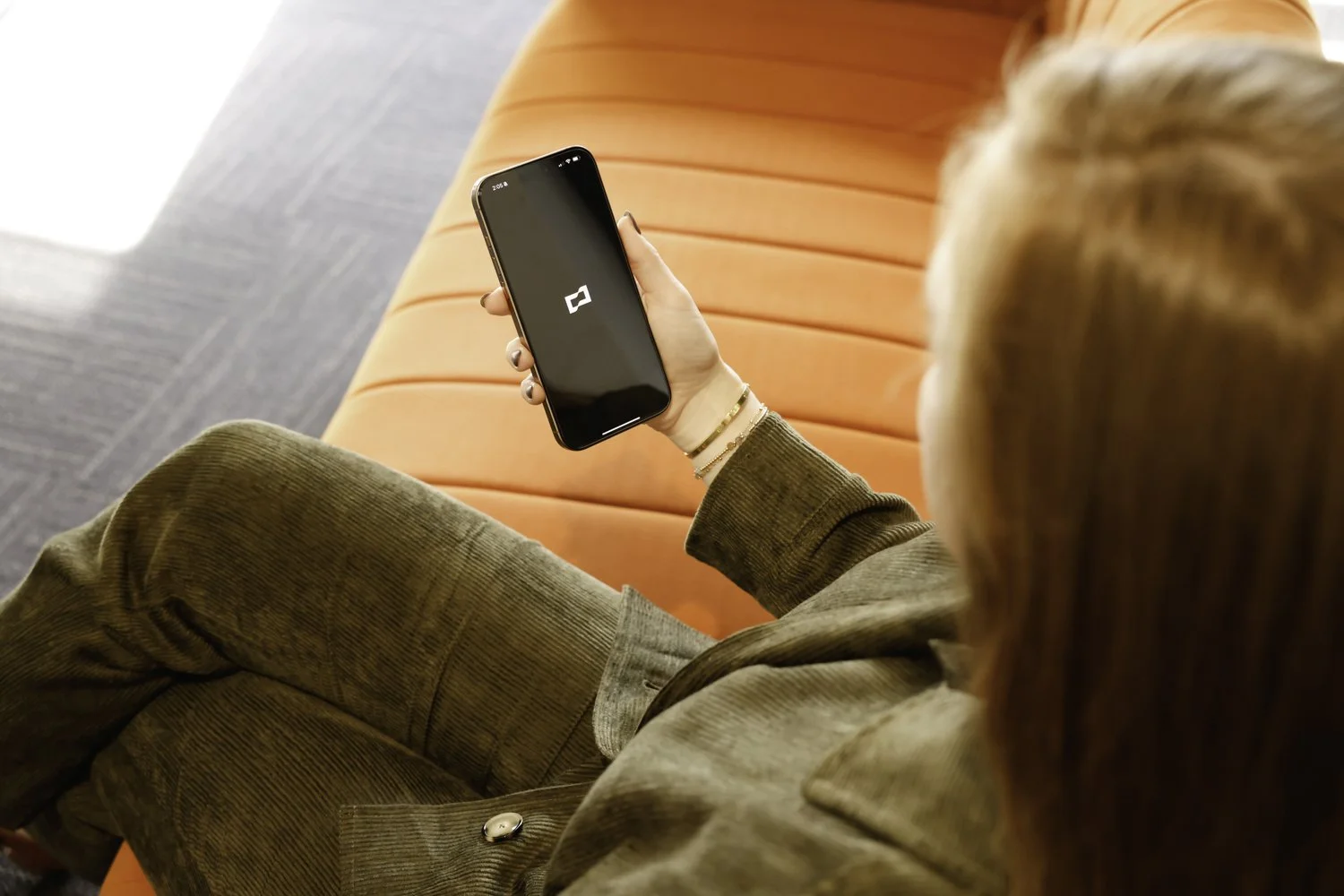 A woman with red hair holding a smartphone displaying an Brex logo, sitting on an orange sofa at Brex Headquarter in San Francisco, CA.