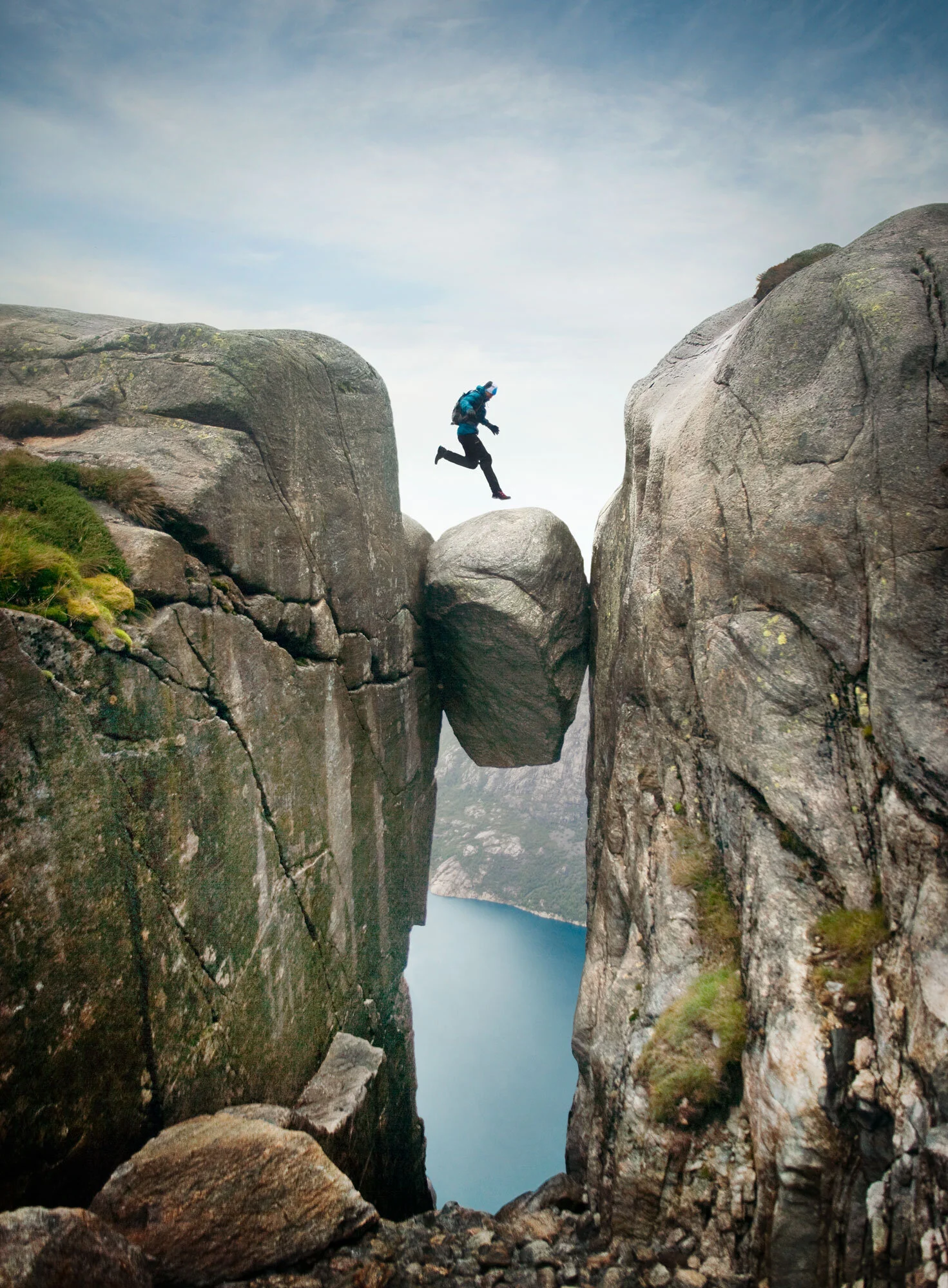 A young man with a backpack jumping over a boulder between two rocks on a mountain, with a body of water below and cloudy weather at Kjeragbolten, Norway.