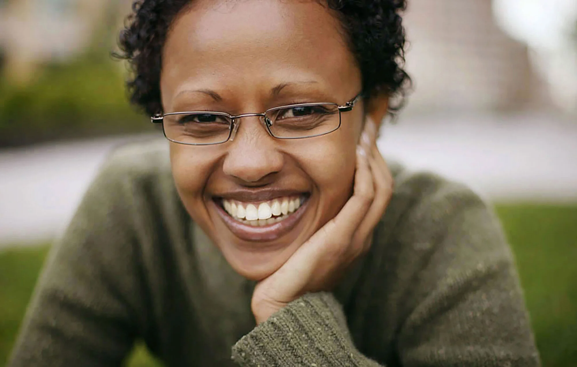 Close-up of a smiling woman with short curly hair, glasses, and a green sweater, resting her chin on her hand outdoors.