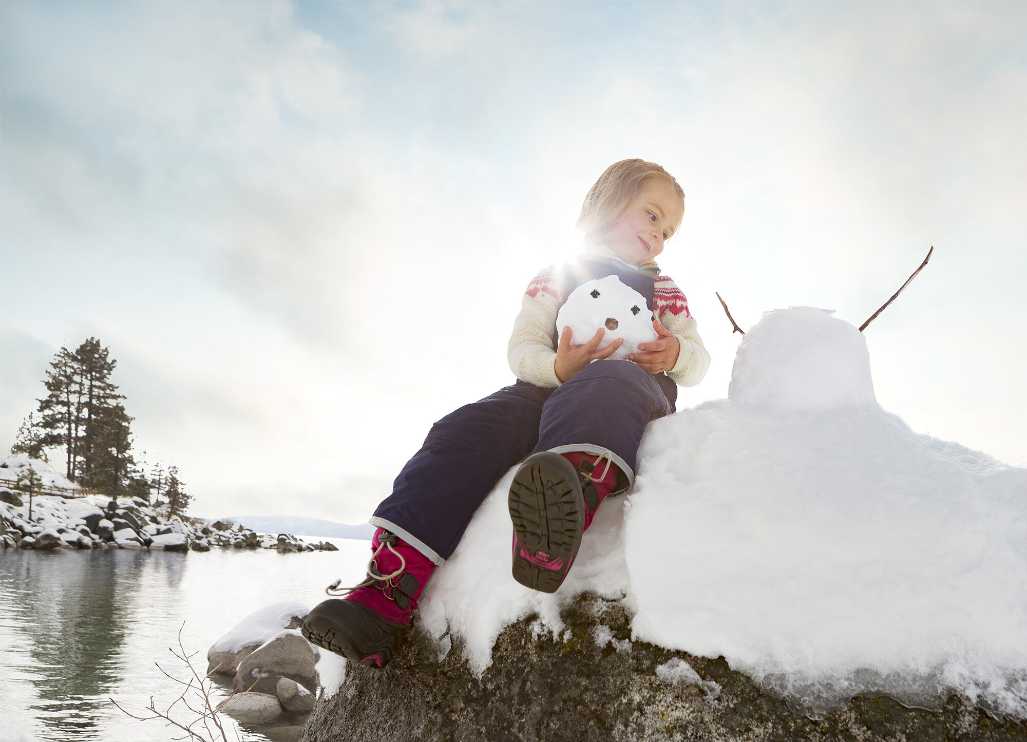 A young girl sitting on a snow-covered rock by a Sand harbor, Tahoe, wearing a wool sweater and skipants, holding a small snowman with a face made of natural materials and a larger snowman with twig arms nearby, under a partly cloudy sky with the sun