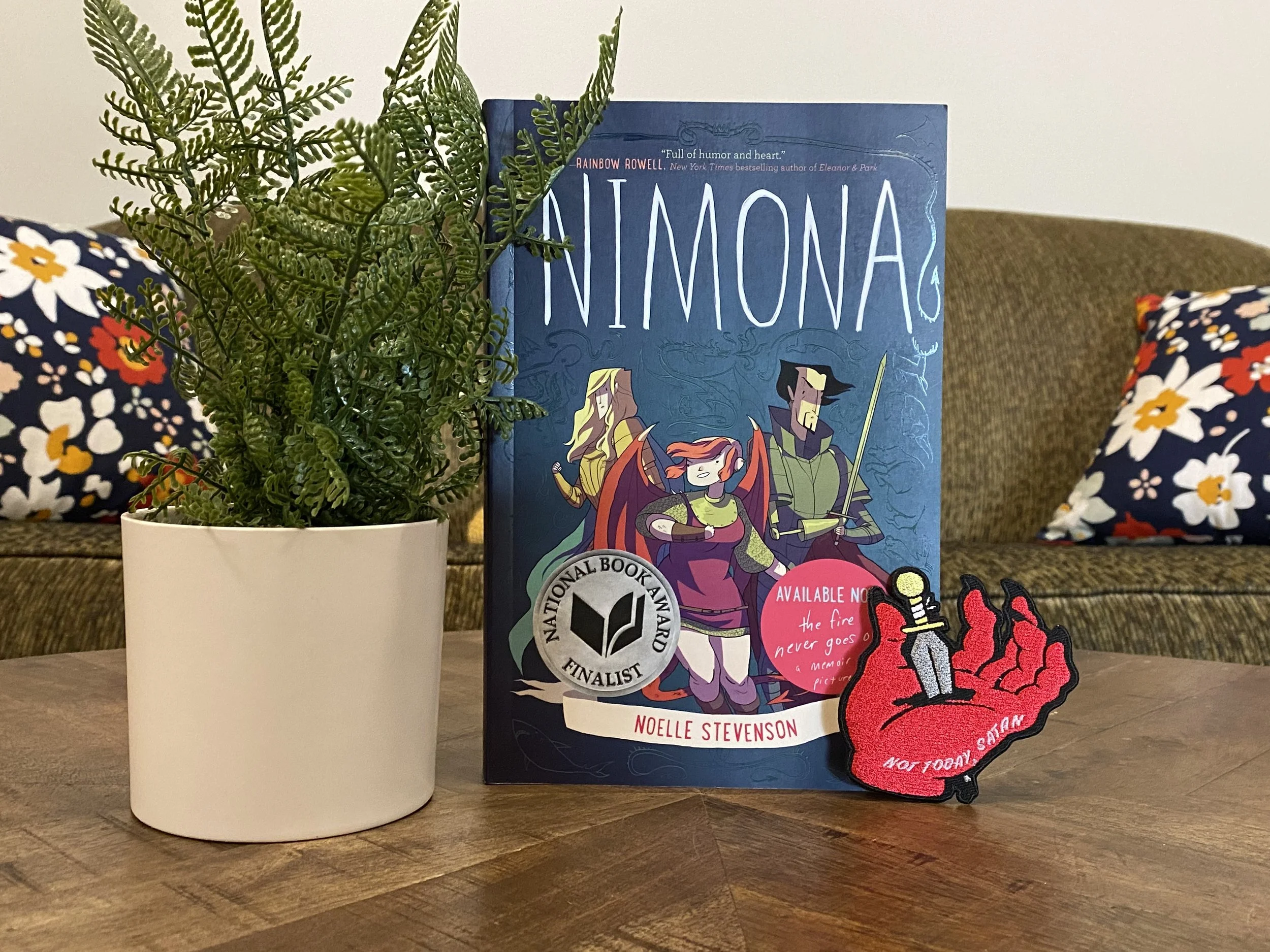 Book Cover. Nimona, young woman smiling evilly with large wings and a tail, stands between a tall man holding a sword with real and artificial hands and a long-haired blond man in golden armor. Next to a patch of a dagger stabbing through a hand.