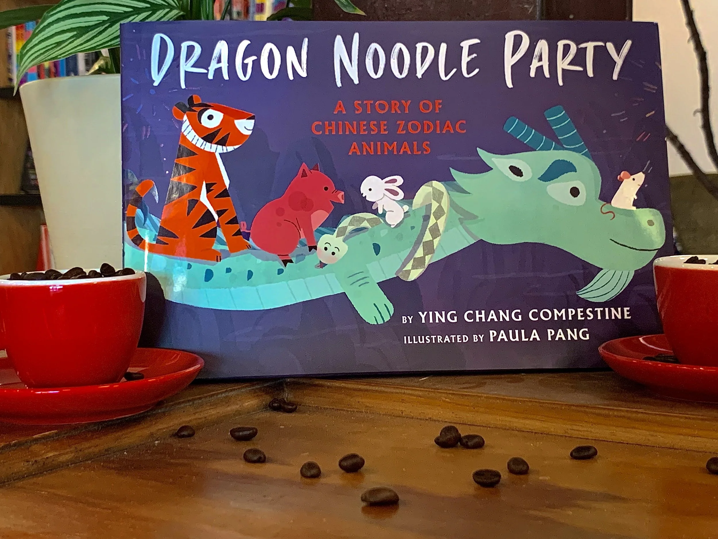 Book cover. A mouse, snake, rabbit, pig, and tiger ride on a dragon's back. Arranged with two red espresso cups filled with beans and a handful of beans scattered on the tabletop.