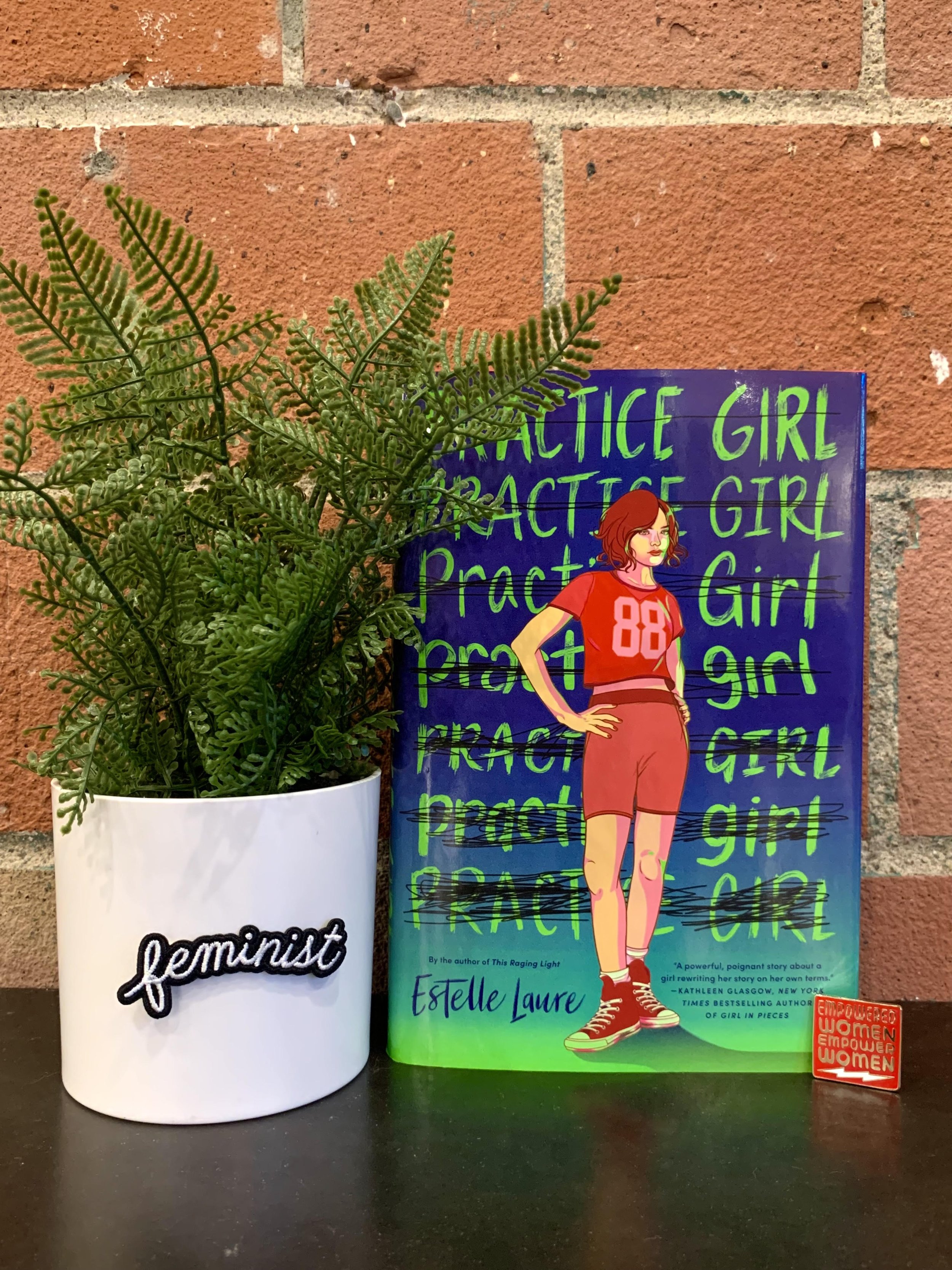 Book cover: a young woman wearing athletic shorts and a jersey rests her hands on her hips. "Practice Girl" is crossed out several times. Arranged with an enamel pin reading "Empowered Women Empower Women" and an embroidered patch reading "feminist."