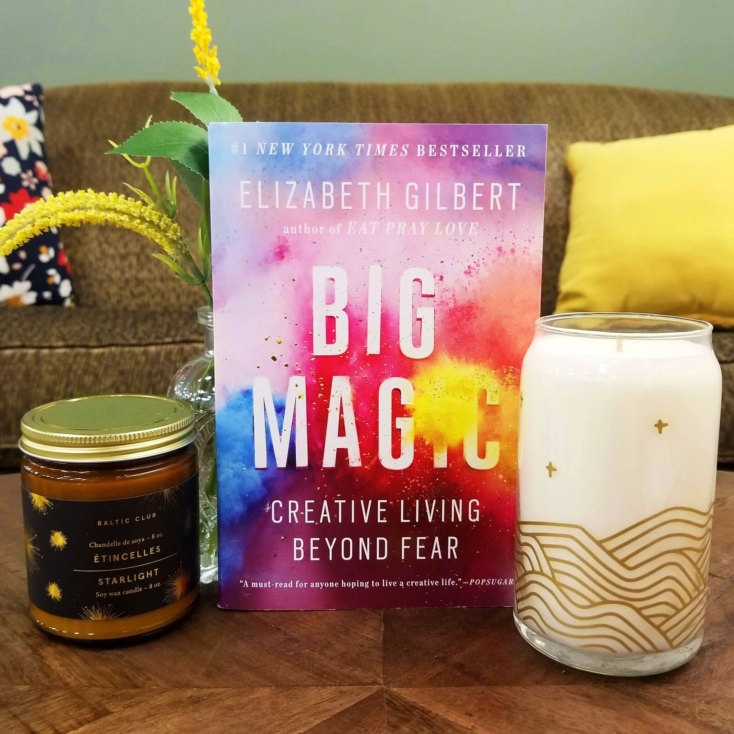 Collage of reviewed book ("Big Magic"),  two glass candles covered in stars, and a vase of flowers. A plush couch with two pillows is in the background.