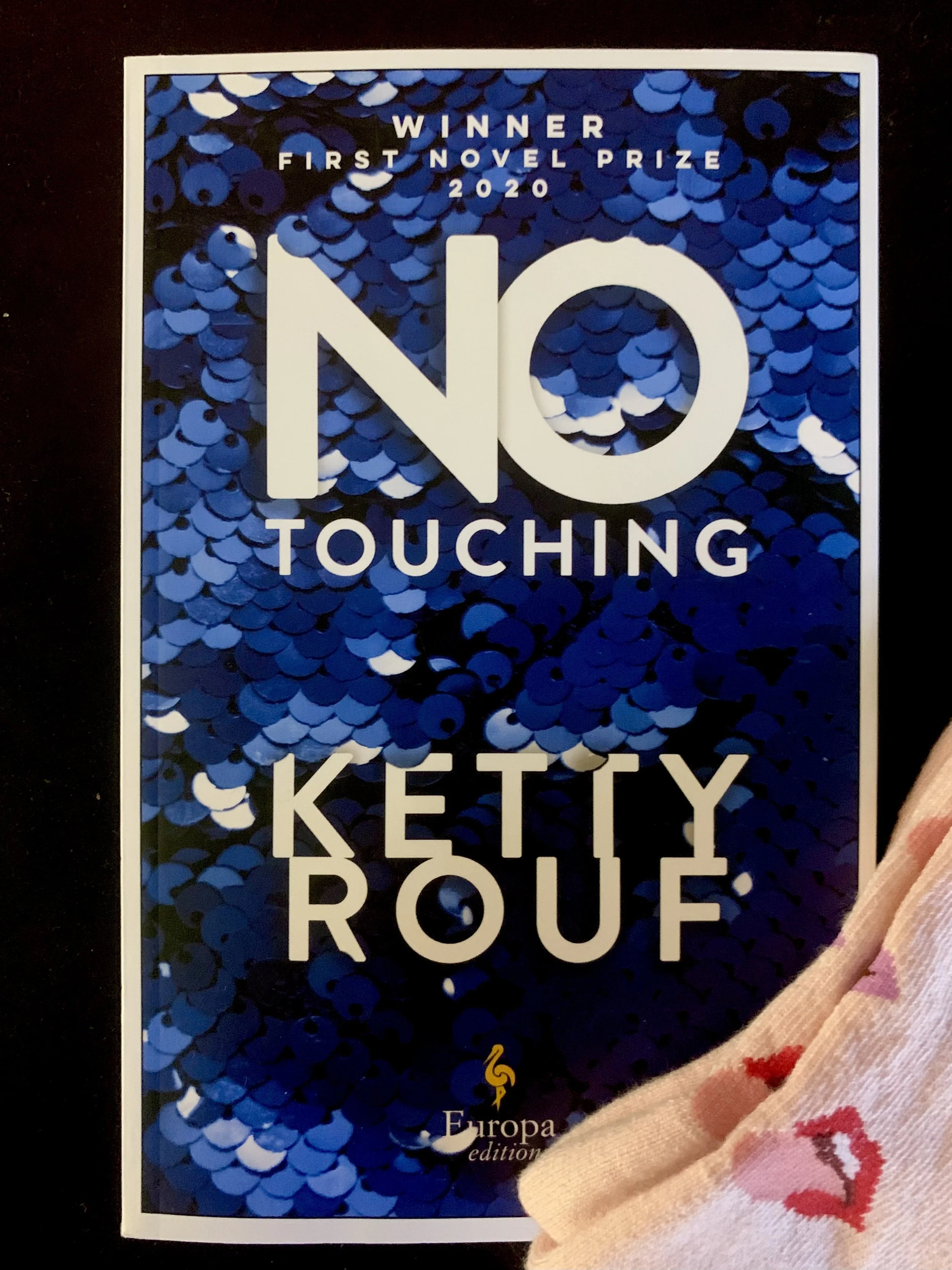 Book cover. Text:  "Winner: First Novel Prize 2020. No Touching. Ketty Rouf" in front of a sequined background. Arranged with a pair of socks covered in mouths sticking out their tongues.