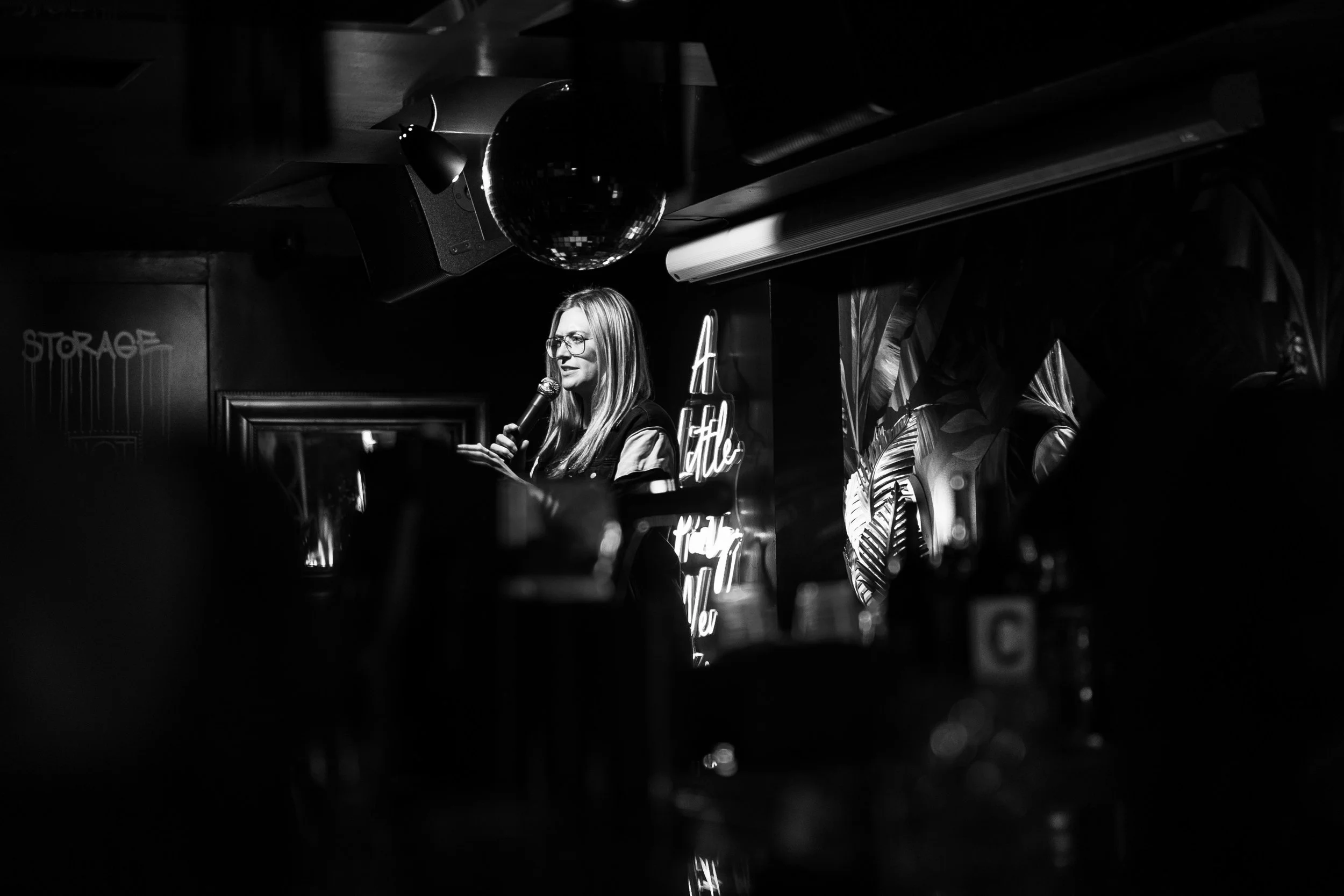A woman with glasses speaking into a microphone on a stage in a dimly lit, black-and-white setting, with neon signs and tropical foliage in the background.