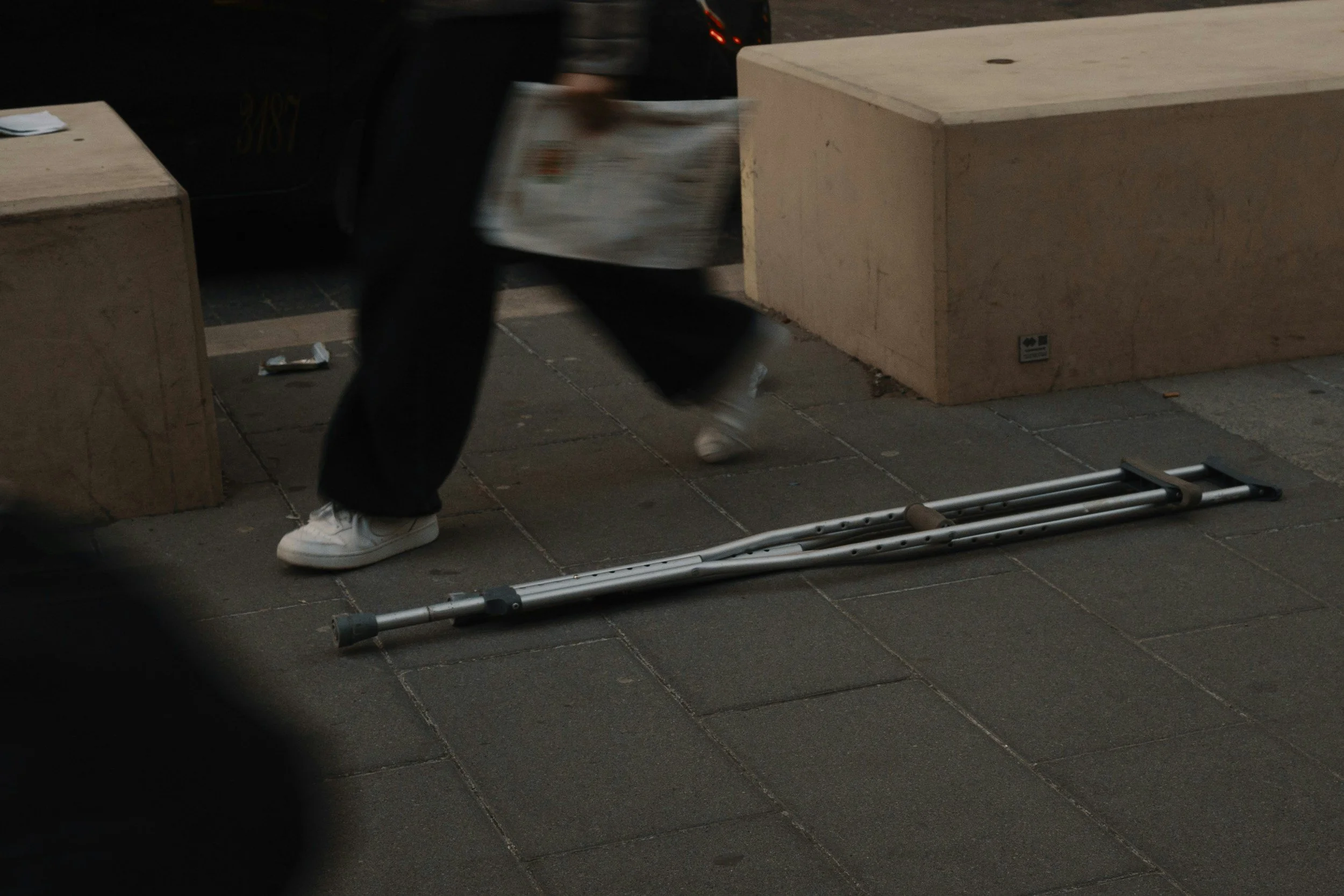 Person walking past two concrete benches on a sidewalk, with a crutch lying on the ground nearby.