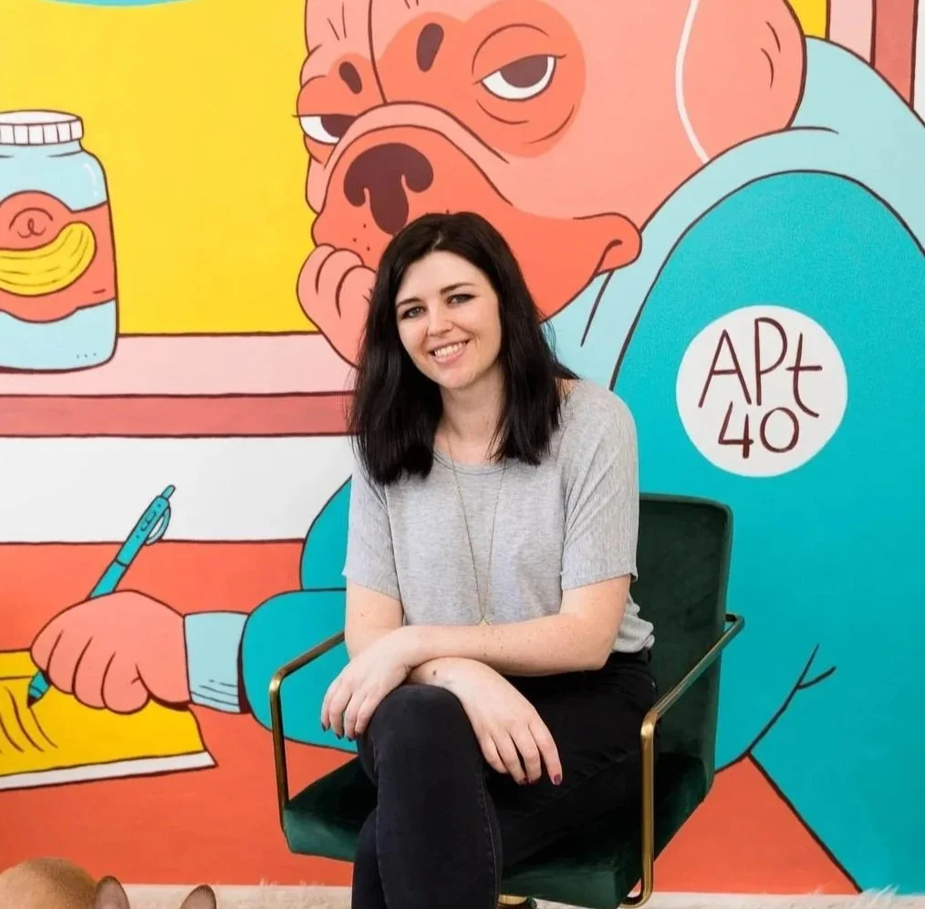 A woman smiling and sitting on a green chair in front of a colorful cartoon mural featuring a large french bulldog holding a pen and a jar of bananas.