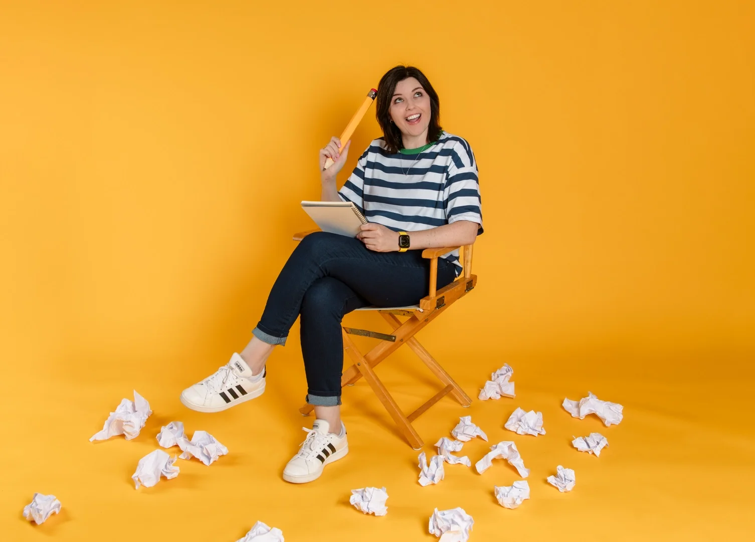 Photo of brand illustrator, Amy, sitting holding giant pencil against yellow background surrounded by scrunched paper balls