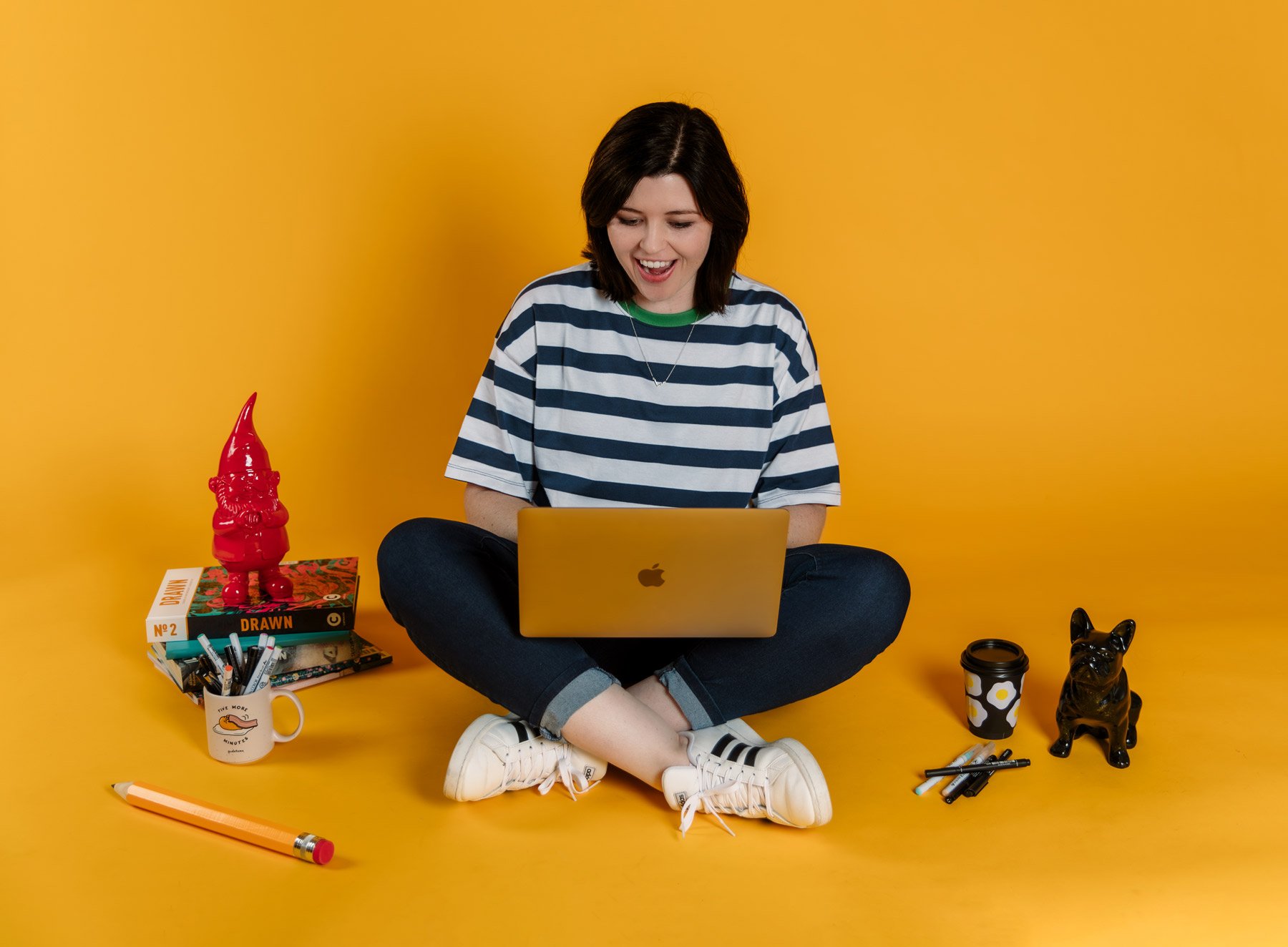 Amy sitting on the floor with a laptop smiling on a yellow background