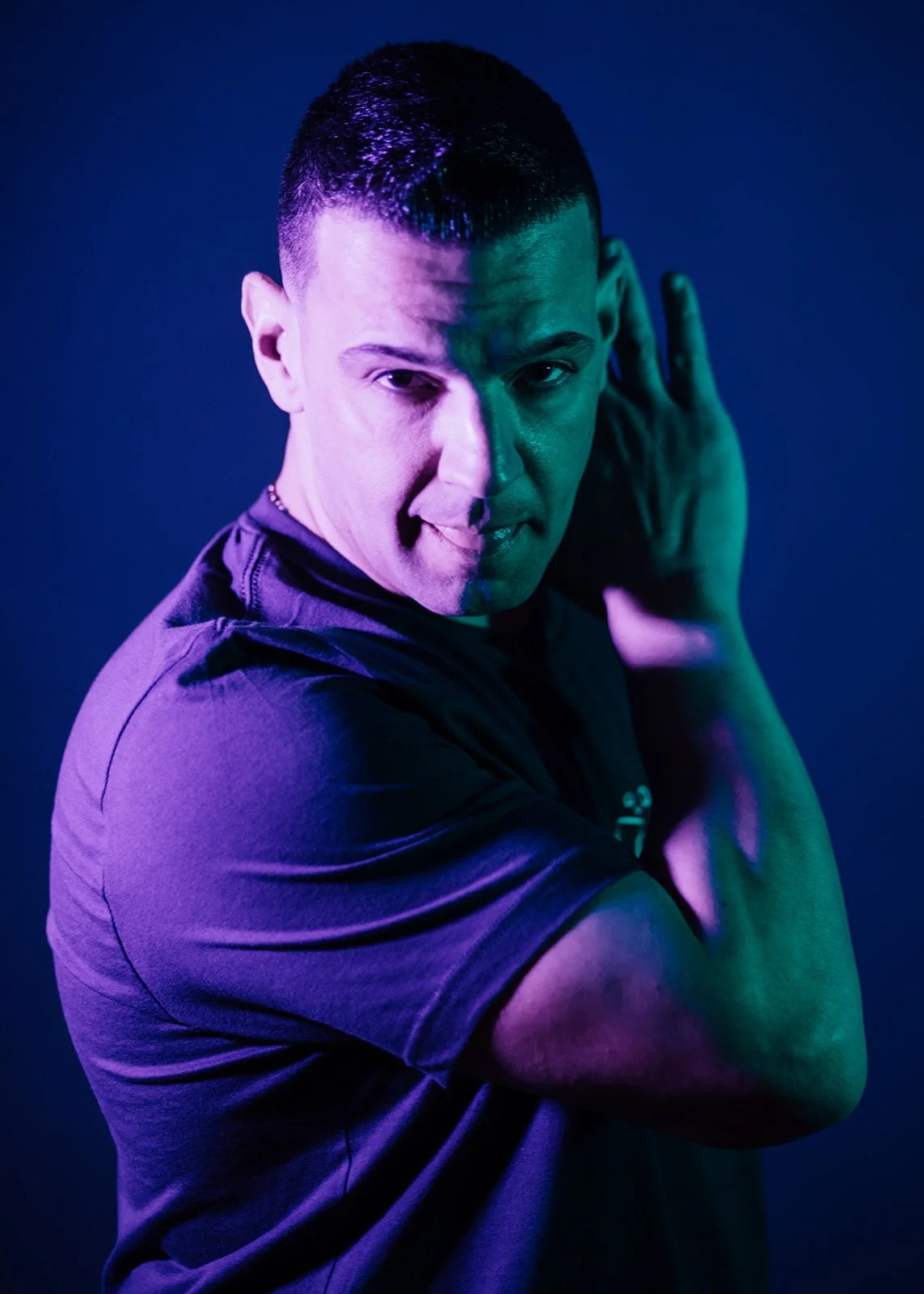 A man with short dark hair, wearing a dark t-shirt, holding his hands near his ears, with dramatic colored lighting on his face, half purple and half green.