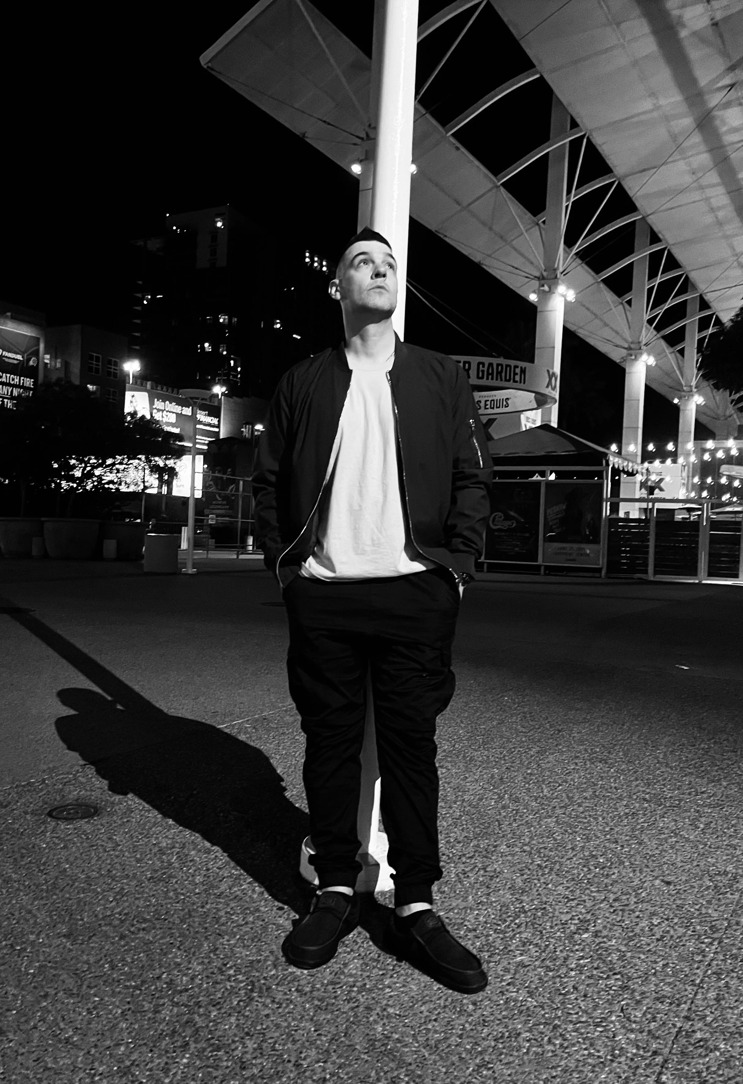 A man in a black jacket and pants standing with hands in pockets at night near a streetlamp, with a modern structure and city lights in the background.